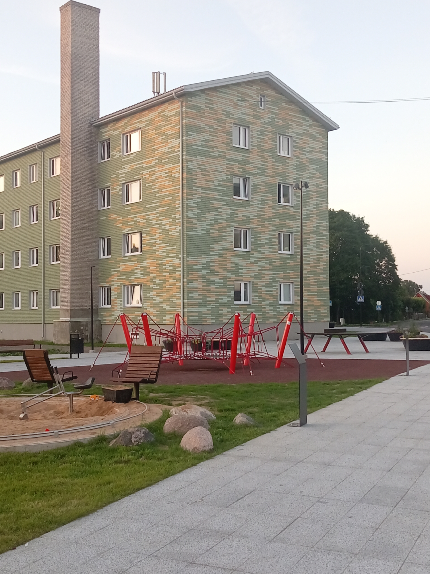 The image depicts a modern apartment building with a brick chimney in Tapa, Estonia. The building features a colorful facade of green, orange, and beige bricks, with numerous white-framed windows. In front of the building, a playground area is visible, including a red net climbing structure, two wooden benches, a sandbox with a mechanical digger, and a table with red supports. The ground is covered with red rubber mulch in the playground and a paved walkway of grey tiles. The surrounding area includes a patch of grass, large rocks, a trash can, a lamppost, and trees in the background. The sky is clear and light.