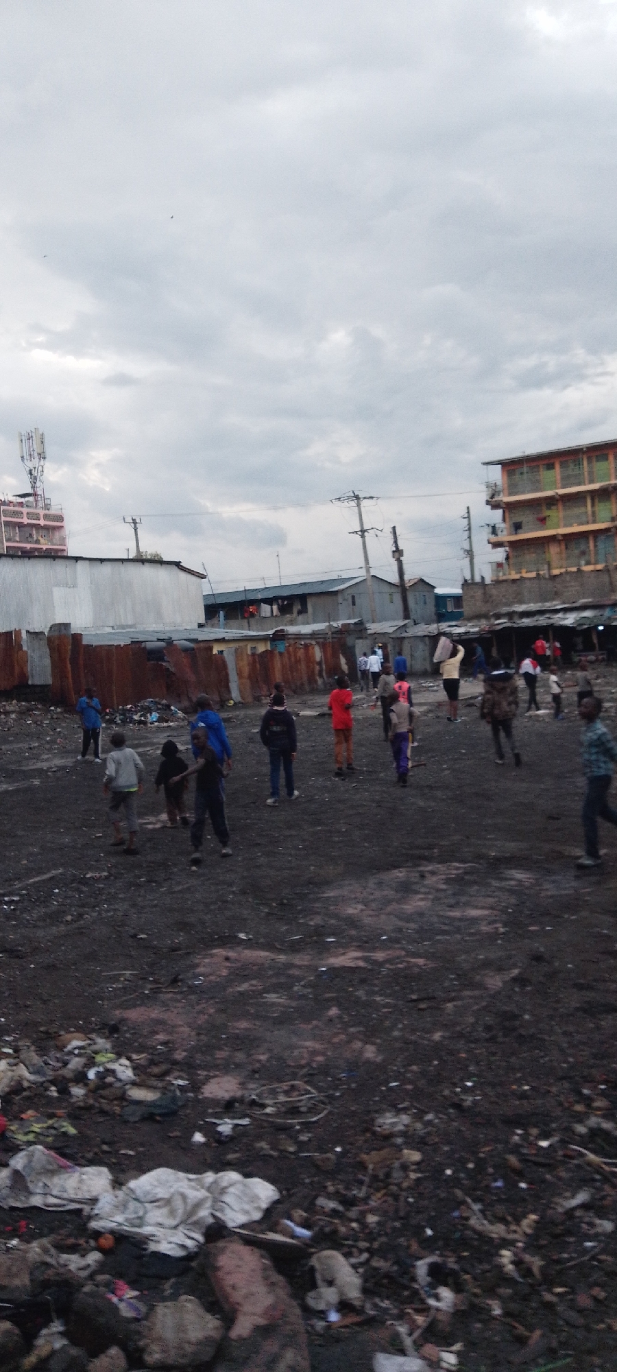 The image depicts a scene in Nairobi, Kenya, characterized by poverty and urban blight. A group of people, including many children, are walking through a large, barren, dirt field covered in trash and debris. Metal shacks and dilapidated buildings surround the field. Power lines are visible overhead. The sky is overcast.