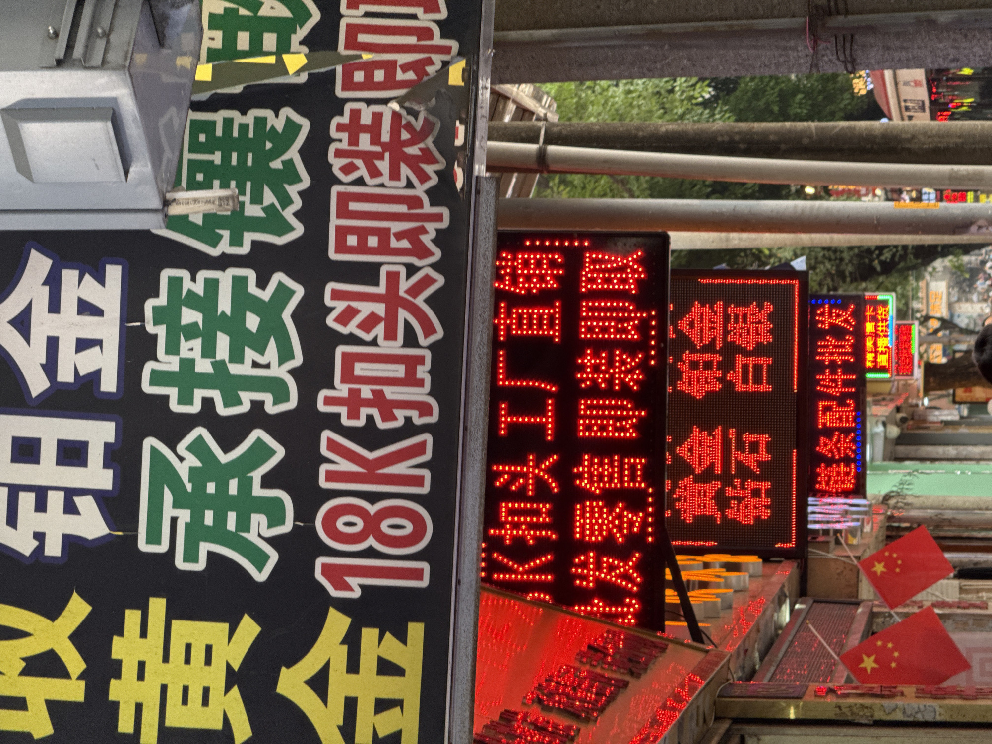 The image depicts a street scene in Liwan District, China, characterized by storefront signs and displays. The sign on the left advertises gold and platinum services, including 18K gold clasps. To the right, LED signs display information about wholesale and retail of gold, platinum, diamonds, and silver. A sign also notes chain parts and wholesale. Chinese flags are displayed in the foreground.