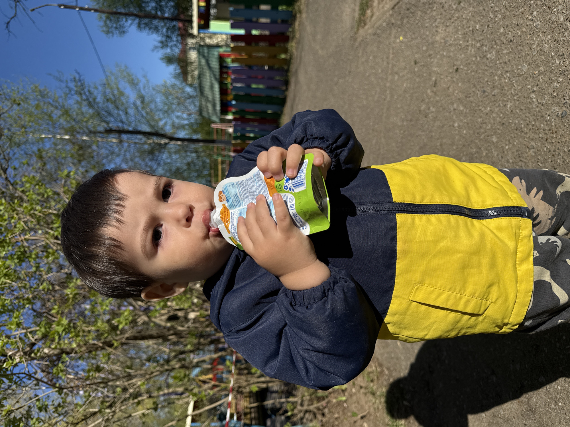 A young child is drinking from a juice pouch outdoors on a sunny day. The child is wearing a dark blue jacket with yellow panels and camouflage pants. The pouch is green and white with an image of a cartoon character. In the background, there is a colorful wooden fence, trees, and a blue sky. The location is in Chaikovsky, Russia. The ground appears to be a paved walkway.