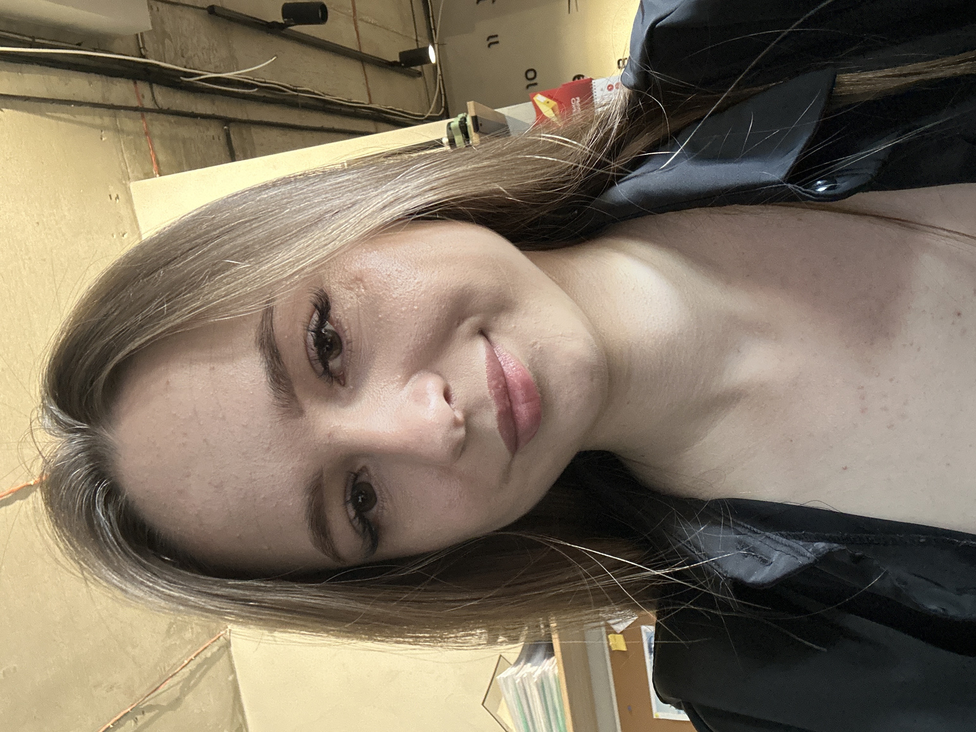 A medium shot shows a young woman with light brown hair and fair skin smiling gently. She is wearing a black button-up shirt. Her hair is down and falls around her face and shoulders. The background appears to be an indoor environment, possibly an office, with a textured ceiling and exposed pipes and wiring. There are shelves with books and papers on the left side of the frame, and a calendar with numbers and a yellow graphic is visible on the right. The lighting is natural and casts soft shadows.
