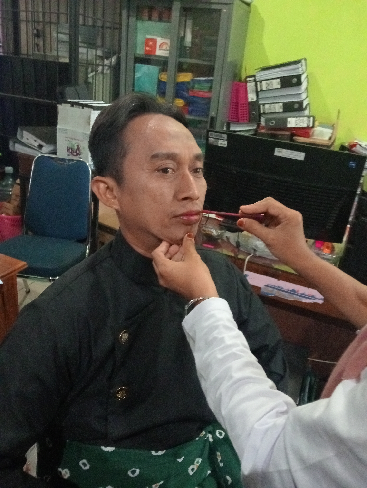 In Demak, Indonesia, an individual is having their lipstick applied by another person. The individual receiving the makeup application is a man with short, dark hair, wearing a black buttoned shirt and a green patterned sash. The person applying the makeup is wearing a white shirt and has their hands visible, one holding a lipstick applicator near the man's lips. The setting appears to be an office or a room with furniture, including a desk, chairs, and shelves filled with binders and other items. The wall is painted lime green.