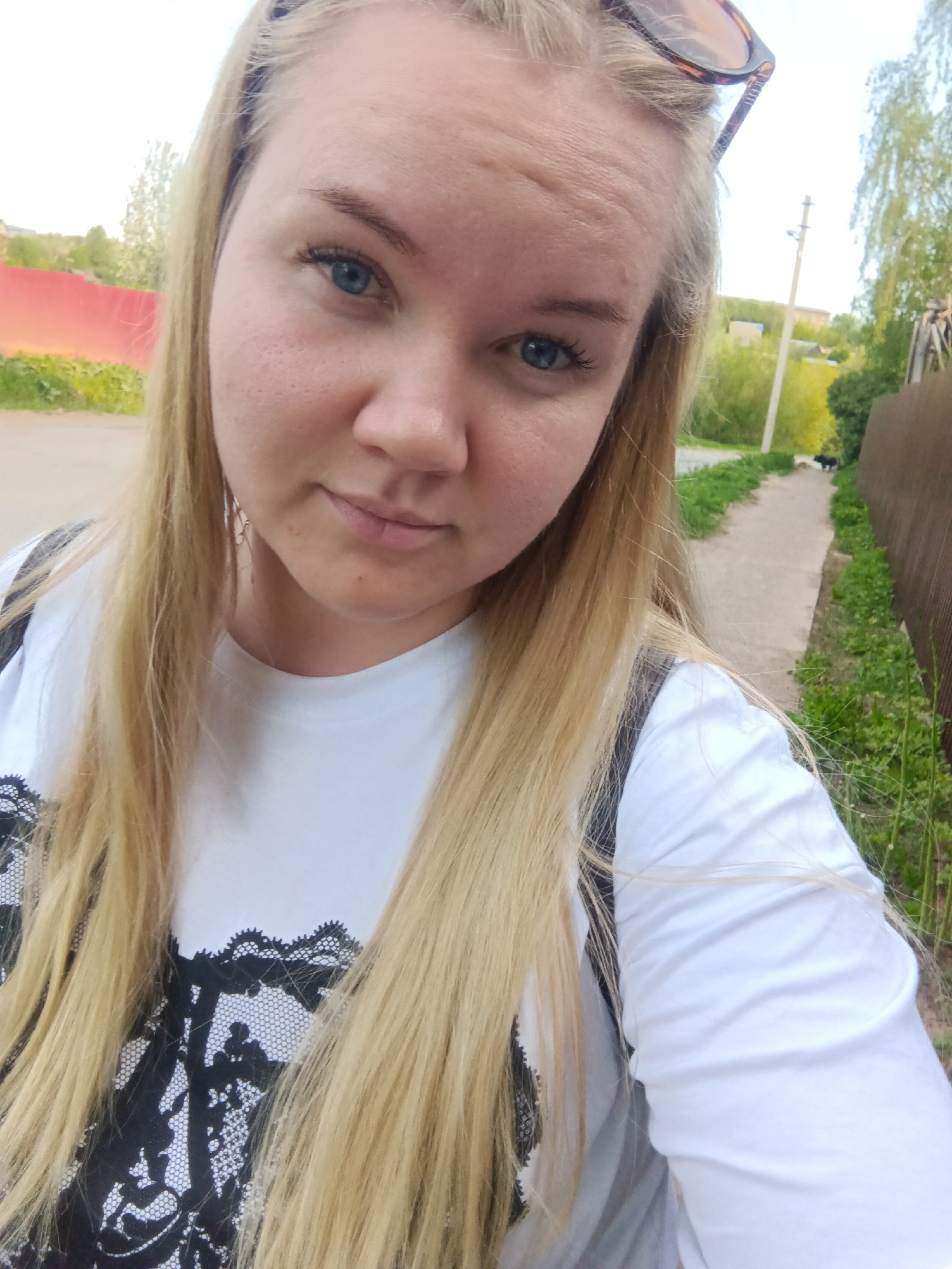 A woman with blonde hair and blue eyes is taking a selfie. She is wearing a white t-shirt with a black lace design. Sunglasses are perched on her head. The background suggests an outdoor location, possibly a residential area. A concrete path bordered by green vegetation and a brown wooden fence can be seen to the right of the frame. Further in the background, there are trees and a utility pole. Given the Фоки, Russia tag, it can be assumed that the image was taken in or near that city.