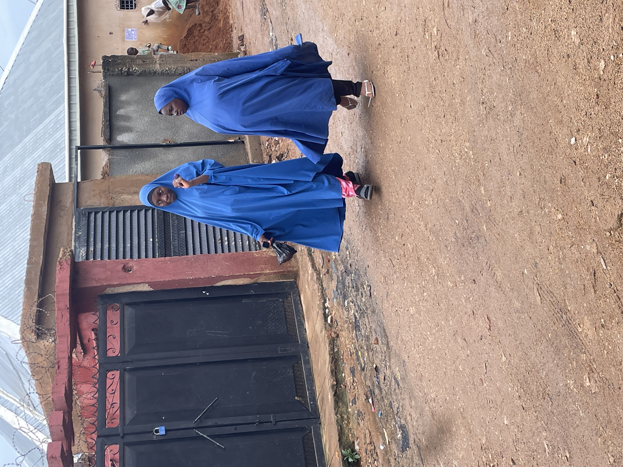 In Gura Top, Nigeria, two women walk on a dirt road. They are both wearing blue hijabs. The woman on the left is holding a black bag. The woman on the right is walking behind her. There are buildings and fences in the background. The road is muddy and there is some debris on it.
