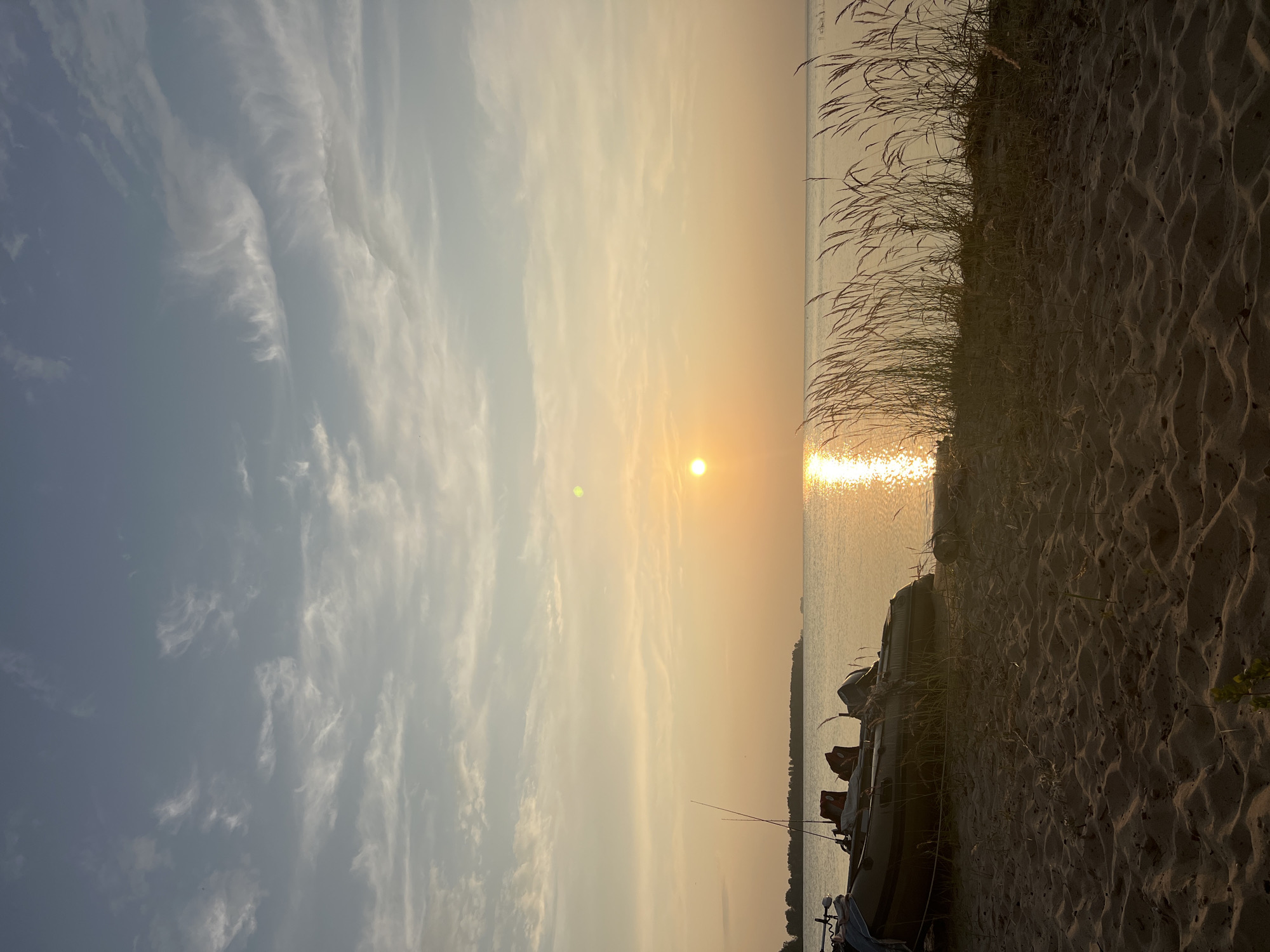 The image depicts a serene sunset scene on a sandy beach, with the calm waters of a lake stretching towards a distant tree-lined horizon. A small inflatable boat with two outboard motors is partially visible on the sand in the foreground, with fishing rods attached. Tall, dry grass lines the edge of the water. The sky is a pale blue with wispy white clouds, gradually transitioning to a warm golden hue near the setting sun, which casts a shimmering reflection on the water. The overall atmosphere is peaceful and tranquil, suggesting a quiet evening by the water. The provided location is Borovoe, Russia, which is not a city but a resort town in Kazakhstan.