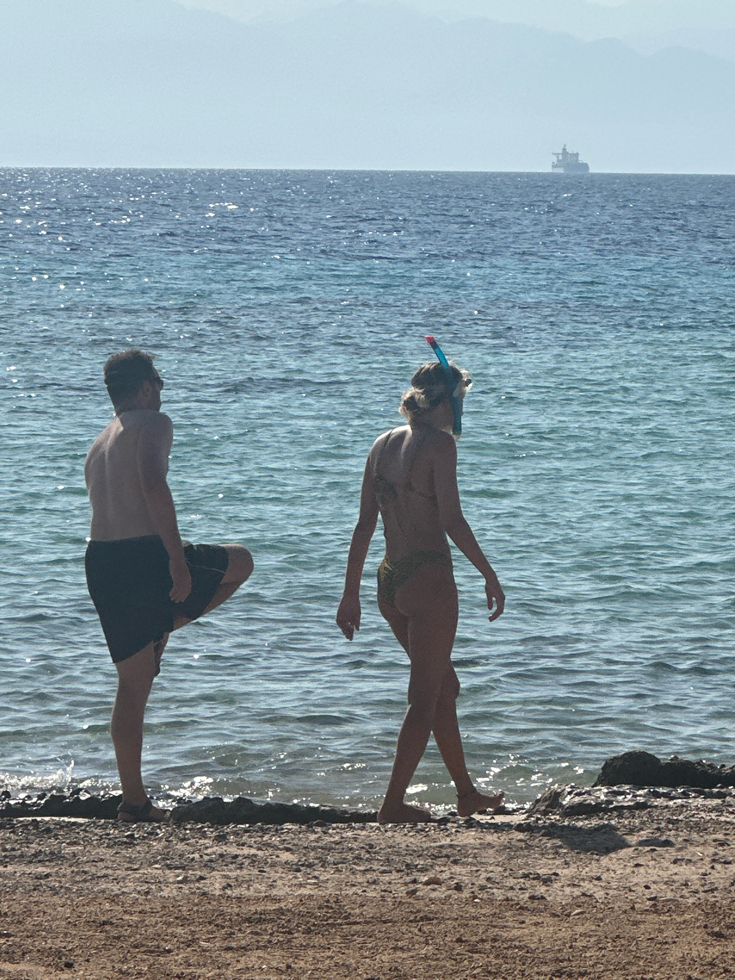 An outdoor, full-shot photograph shows two individuals standing at the water's edge on a sandy and rocky shoreline. The water, a deep blue with shimmering highlights from the sun, stretches to the horizon where a faint, hazy mountain range is visible. A large cargo ship is positioned on the horizon, centered in the distance. The person on the left, a bare-chested male wearing dark swim trunks, stands with his right leg bent and lifted. The person on the right, a female wearing a bikini and snorkeling gear, is facing away from the viewer, looking towards the water. Her hair is pulled back, and she is wearing a snorkel mask and tube. The shoreline is composed of light brown sand mixed with dark pebbles and rocks. The lighting suggests it is daytime, likely mid-day due to the bright sunlight. The location is the Red Sea coast in Nueiba, Egypt.