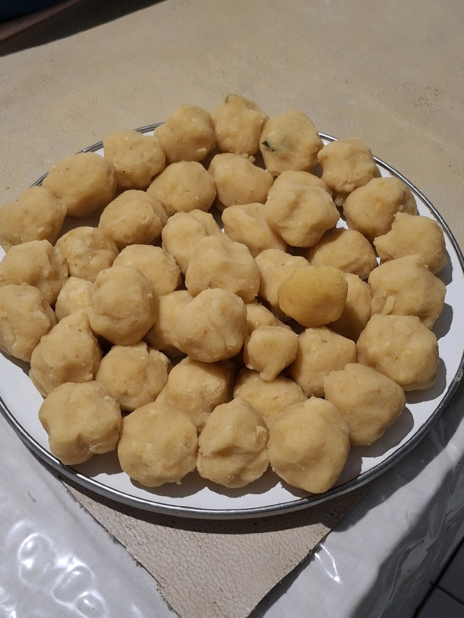 A close-up shot displays a plate filled with numerous small, golden-brown food items, possibly potato fritters or dumplings, arranged in multiple layers. The plate has a white rim with a thin silver band. The food items are spherical to slightly irregular in shape, with a somewhat lumpy texture, suggesting they are homemade. Some items have subtle variations in color and may contain small green flecks. The plate rests on a light-colored, textured surface that could be a table or countertop. In the background, a darker surface is visible, possibly a wall or another piece of furniture. The lighting creates shadows around the food items, highlighting their three-dimensional form. The image captures a food preparation or serving scenario, likely in a domestic setting within Lumajang, Indonesia.