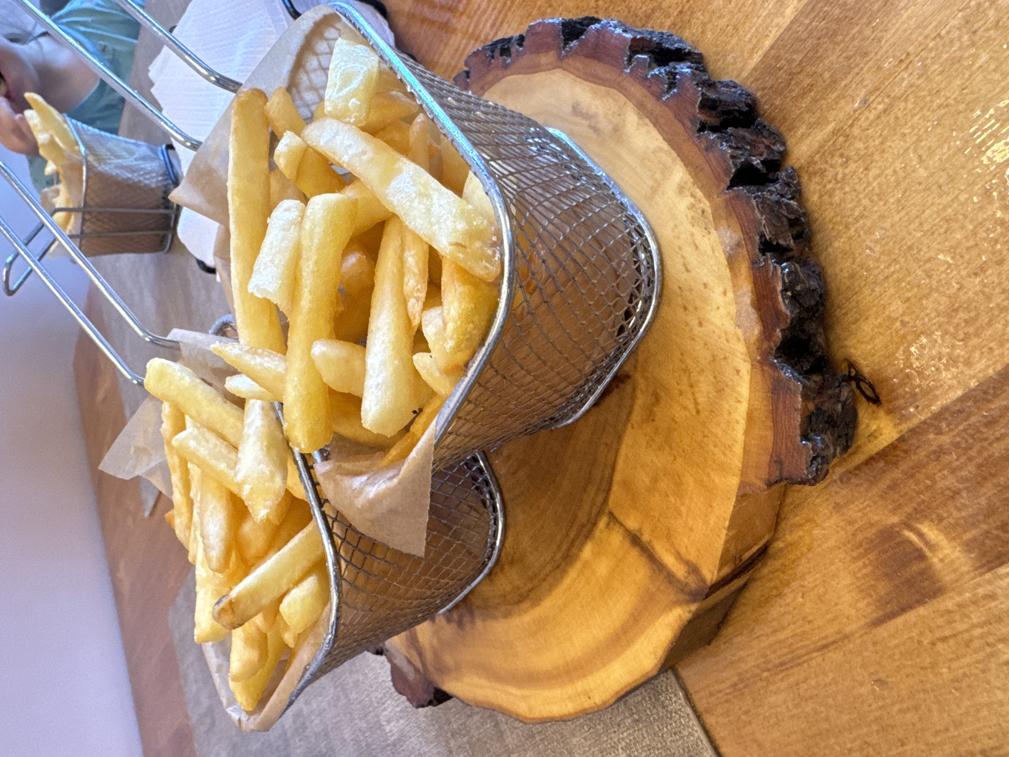 In a close-up, high-angle shot, two wire baskets filled with golden French fries are presented on a rustic wooden slab. The fries are uniformly cut and appear to be freshly fried, with some showing slight browning. The baskets are lined with brown paper, and one is slightly tilted, allowing some fries to spill over. The wooden slab has a natural, uneven edge with dark bark, showcasing the wood grain. In the background, a person wearing a light blue shirt is partially visible, with their hand reaching towards the fries. The lighting suggests an indoor setting, possibly a restaurant or dining area. The scene evokes a sense of casual dining and enjoyment.