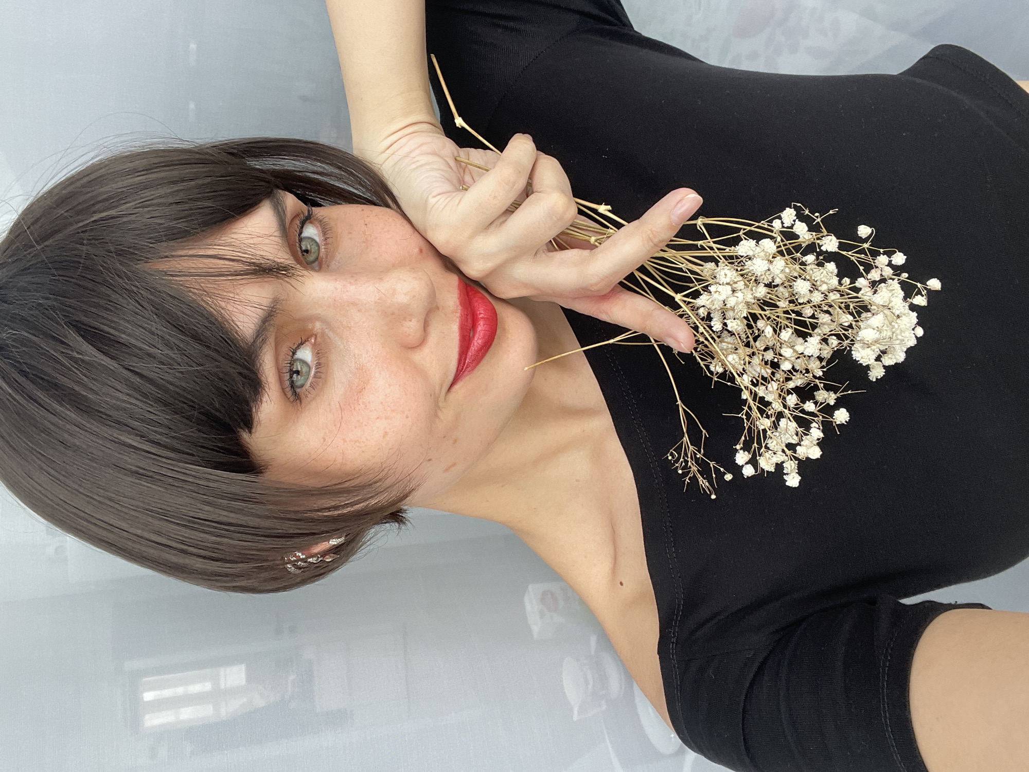 A woman with short, dark hair and green eyes is holding a bouquet of dried white flowers. She is wearing a black top and red lipstick. Her left hand is touching her face. The background is a light grey surface, possibly a wall or a floor. There is a reflection of a window or light source visible on the surface. There is no indication of this image being taken in Izhevsk, Russia.