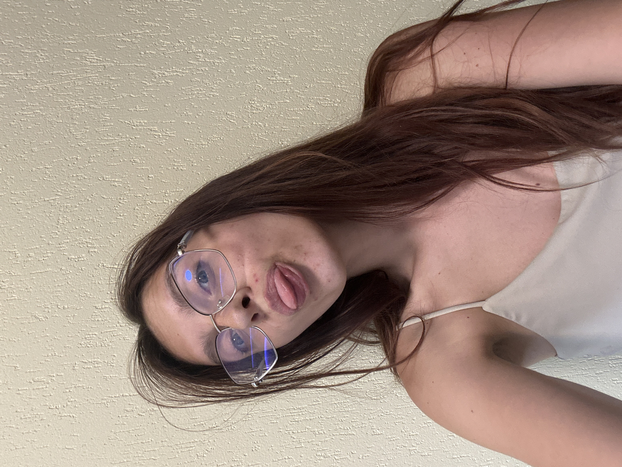 A young woman with long, dark brown hair and light blue eyes is pictured from the chest up, looking at the camera. She is wearing large, clear-framed glasses and sticking her tongue out slightly. Her skin has freckles and some minor blemishes. She is wearing a light-colored spaghetti-strap top. The background is a textured, light-colored wall. The image appears to be a selfie taken indoors. The location of Tryavna, Bulgaria, is not indicated or discernible from the image.