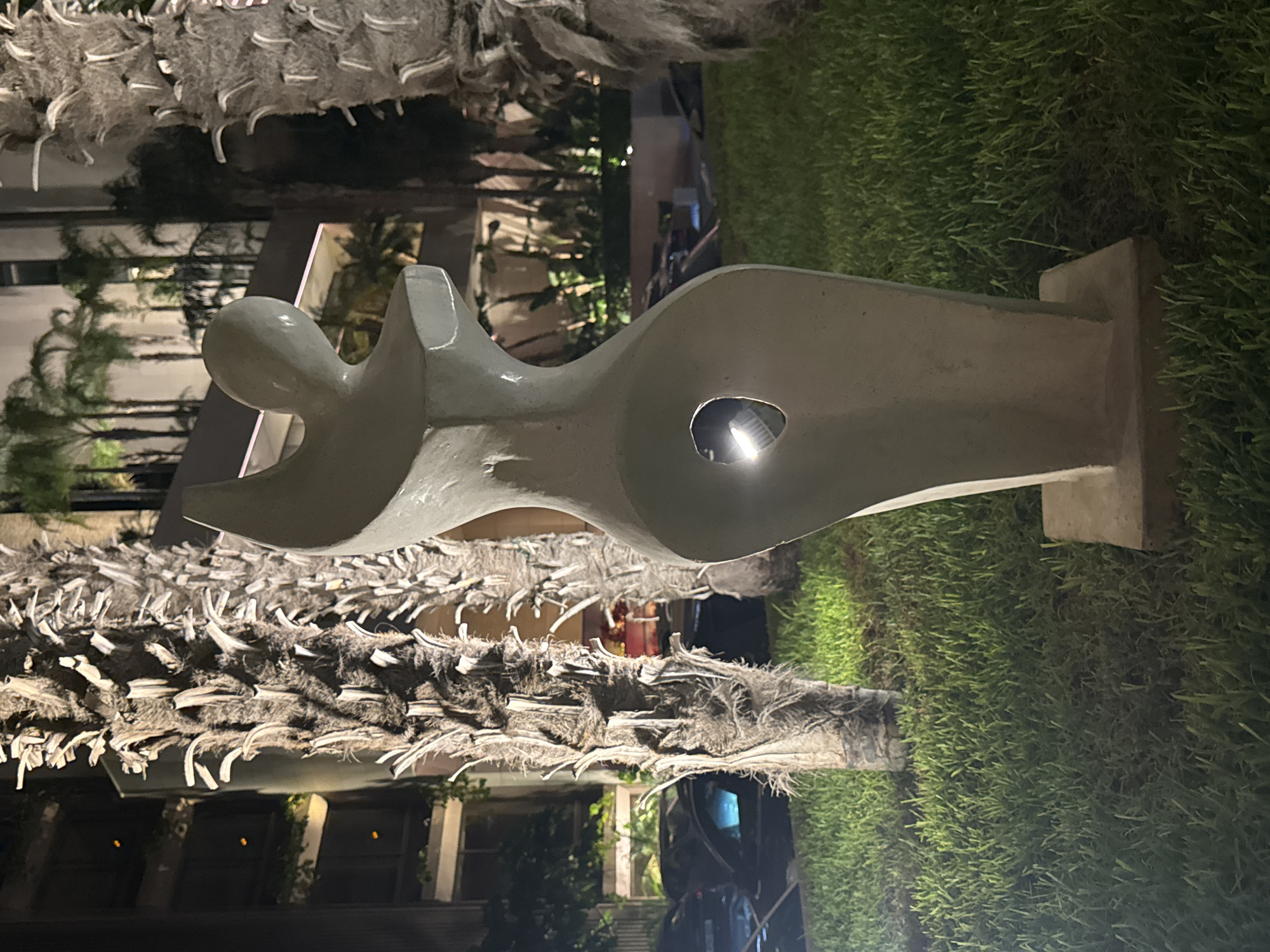 A nighttime exterior shot shows a light-colored abstract sculpture with a rounded, human-like form, featuring an oval opening in its midsection with a light source visible within, positioned on a concrete base on a manicured lawn. The sculpture is situated between two palm trees with rough, fibrous trunks. In the background, a modern building with illuminated windows and palm trees can be seen. The scene is illuminated by artificial lights, casting shadows and highlighting the textures of the plants and sculpture. The location is identified as Agios Tychona Community, Cyprus.