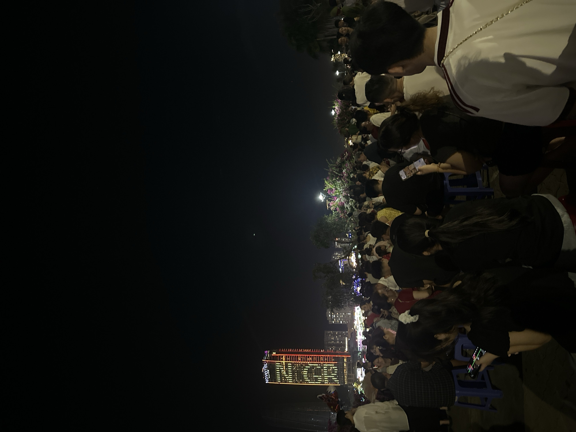 This media file captures a night scene in Đà Nẵng, Vietnam, likely during a festive event. A large crowd of people are gathered, facing towards a brightly lit building that appears to be a hotel, as indicated by the visible text "NOVOTEL" on its facade. The building is adorned with colorful lights, forming patterns and illuminating the night sky.
The subjects are primarily people of various ages, many of whom are looking towards the illuminated building, suggesting they are watching a display or an event. Some individuals are holding up their phones, possibly recording or taking pictures. The atmosphere appears to be one of communal gathering and observation, with a sense of anticipation or enjoyment. The scene is set at night, with the darkness contrasting with the artificial lights from the building and distant cityscapes. Some trees and decorative lights are also visible, adding to the festive ambiance. The weather appears to be clear, as there are no indications of rain or inclement conditions. The crowd density suggests a popular event, drawing a significant number of spectators. The people are dressed in casual attire, consistent with an outdoor evening gathering.