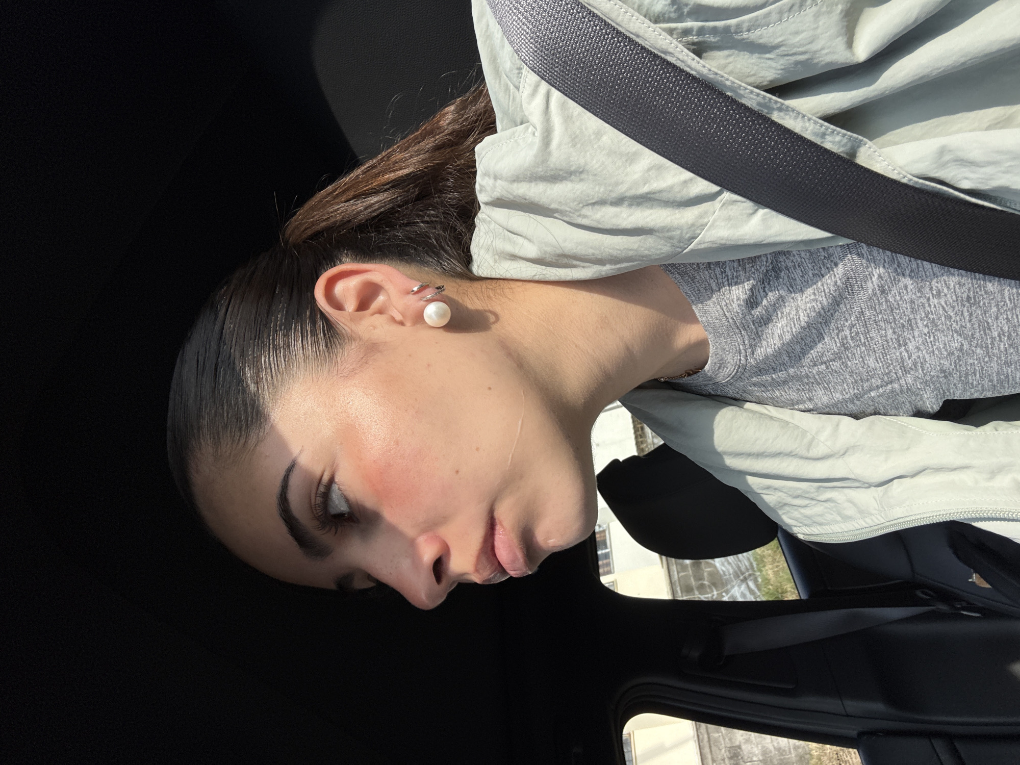 A close-up, chest-up shot features a person, likely a woman, seated inside a car during bright daylight. Her dark hair is pulled back, revealing a large white pearl earring and two smaller silver helix earrings on her left ear. She is wearing a light greenish-grey zip-up jacket over a grey t-shirt, with a dark seatbelt fastened across her chest. Her face, seen in profile looking downwards and to the left, is illuminated by strong sunlight from the right, creating highlights and shadows that emphasize her facial structure, defined eyebrows, and a hint of reddish hue on her cheekbones. A faint line or mark is visible on her left cheek. Her expression appears neutral or pensive. The interior of the car is dark, framing the subject. Through the car window on the right, a blurred background shows light-colored ground, possibly paved, and distant, indistinct structures, suggesting an outdoor location in San Bernardino Tlaxcalancingo, Mexico. No text is visible in the image.