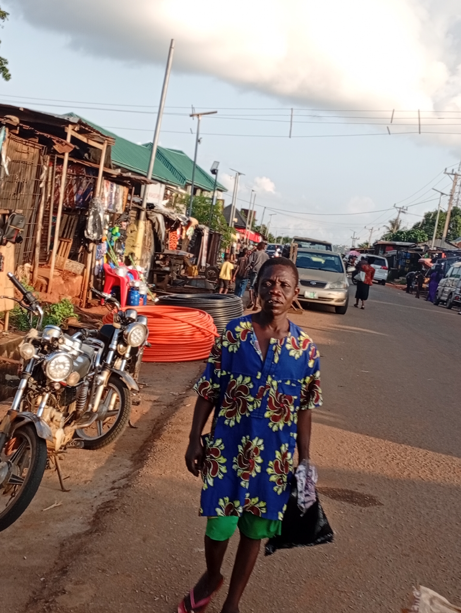 The image depicts a street scene in the city of Nsugbe, Nigeria. In the foreground, a person is walking towards the camera, wearing a blue and red floral print shirt and green shorts. They are carrying a black plastic bag and a white cloth in their right hand. To the left of the person, two motorcycles are parked on the side of the road. Behind the motorcycles, a market stall is visible with various items for sale. The street is lined with more market stalls and buildings. Several vehicles, including a silver car and a white van, are driving on the road. The sky is partly cloudy. The ground is a mixture of dirt and asphalt. The overall impression is of a busy marketplace in a Nigerian city.