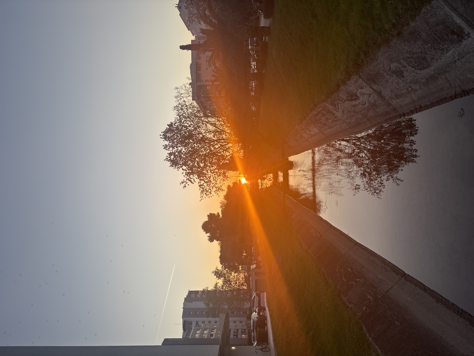 The image depicts a canal or narrow waterway in an urban setting. The sun, partially obscured by trees, casts a bright orange glow across the water and surrounding areas, indicative of either sunrise or sunset. Concrete embankments line the canal, with grass growing adjacent to them. Buildings of varying heights are visible in the background, suggesting an urban residential area. A white contrail from an airplane crosses the clear sky. The sun's glare makes it difficult to discern details, but cars and possibly bicycles are parked along the street next to the canal. There are no people immediately visible in the frame. The image's orientation is rotated 90 degrees clockwise. Location in the city of Bar, Montenegro is not confirmed.