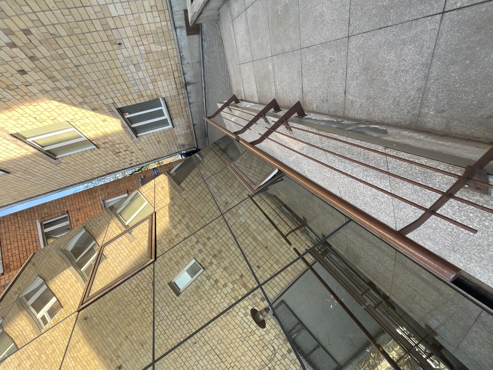 This image depicts the exterior of two buildings, likely located in an urban area. One building is clad in light tan bricks with several rectangular windows, while the other, visible at the edge of the frame, has red bricks. A concrete walkway is present. A metal structure with three horizontal bars runs along the edge of the walkway. The image is taken at an unusual angle, looking upwards.