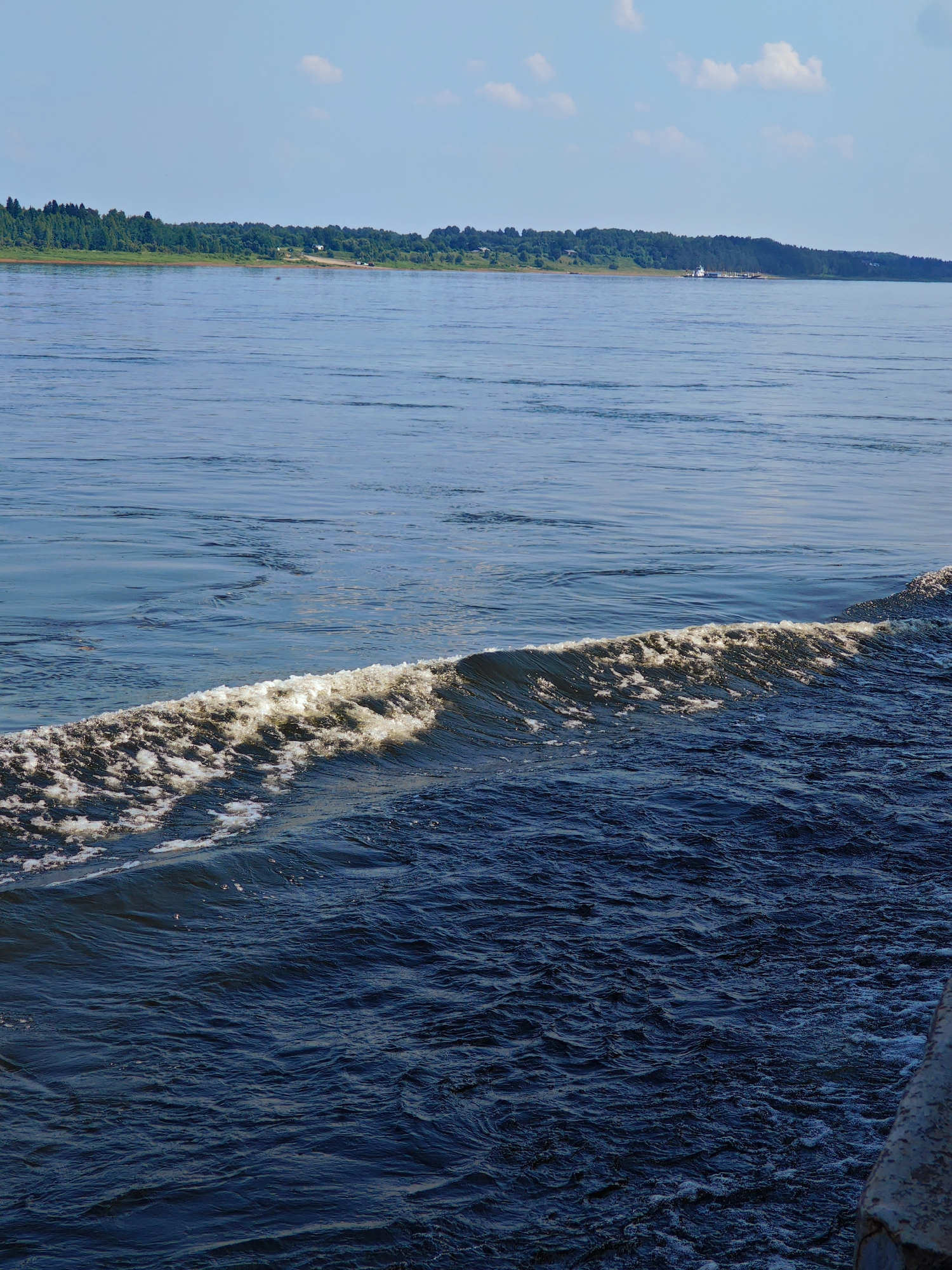 This is a high-angle, full shot of a wide, dark blue river. The river occupies the majority of the frame, with a foamy white wave in the mid-ground and a distant shoreline in the background. The shoreline is lined with a dense forest of green trees and a clear blue sky with a few scattered white clouds above. A small white boat is visible on the water near the distant shore. In the lower right corner of the image, a textured, greyish-brown object, possibly part of a dock or pier, is visible. The scene is devoid of people. The location is the city of Ust'-Kem', Russia.