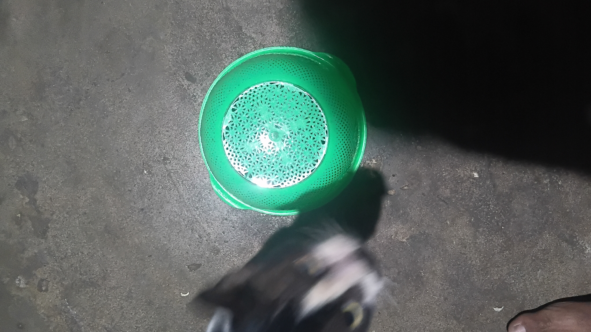 An overhead, slightly angled shot shows a green colander on a dark, textured surface. The colander is translucent and has a decorative, perforated pattern in the center. To the left of the colander, the blurred head of a cat is visible, with its face angled towards the colander. The cat's fur is a mix of black, white, and grey. A human foot, with visible toes and toenail, is partially visible in the bottom left corner of the image, positioned to the left of the cat. The background appears to be a paved or concrete surface, likely outdoors, as indicated by the subtle texture and the presence of ambient light and shadows. The scene is dimly lit, suggesting it was taken at night or in a poorly lit area. The location is consistent with an outdoor setting in a residential or public area within Prabumulih, Indonesia.
