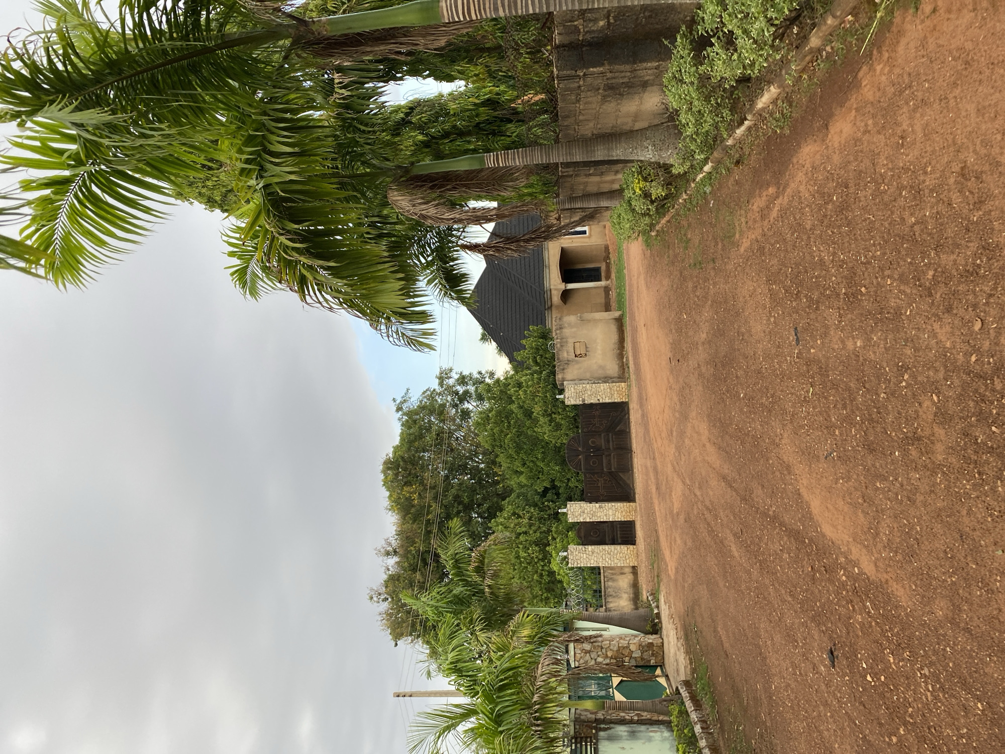 The image shows a dirt road in Bukuru, Nigeria. Palm trees line the left side of the road. On the right, a concrete wall separates the road from a property. A house with a dark roof and light-colored walls is visible further down the road. An ornate metal gate stands at the entrance of the property, surrounded by greenery. The sky is overcast.