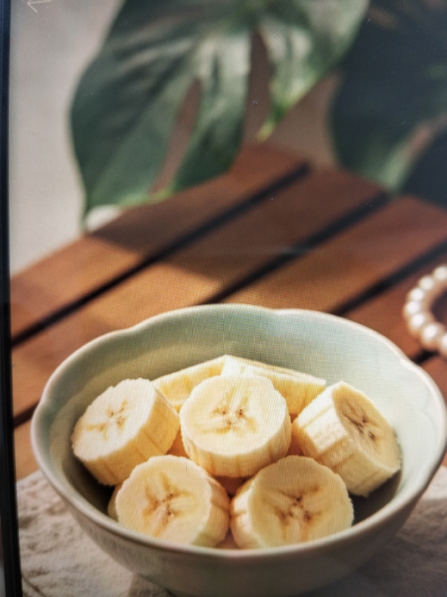 A close-up shot shows sliced bananas in a light teal bowl. The bowl is on a wooden surface, with distinct planks visible. In the background, out of focus, is a large green leaf and what appears to be a string of pearls. The lighting suggests a natural light source, creating shadows on the wooden surface. There are no people present in the image, and no activities are depicted. The image does not provide any information about the city of Payagaboh, Indonesia, or its location.