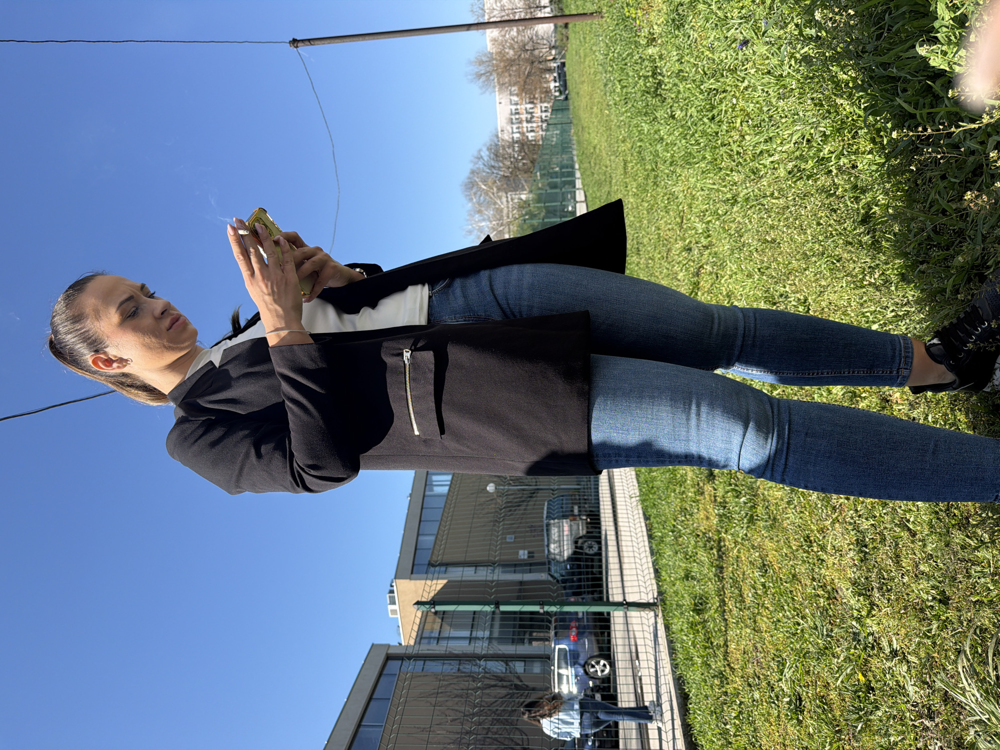 A woman is standing outdoors, likely in a park or grassy area. She is holding a mobile phone in one hand and appears to be smoking a cigarette with the other hand. She is dressed in a black blazer over a white top and jeans. She is looking at her phone. The background shows buildings, trees, and a fence. Given the architectural style and environment, it is likely the city of Plovdiv, Bulgaria, which features a mix of old and modern buildings alongside green spaces.