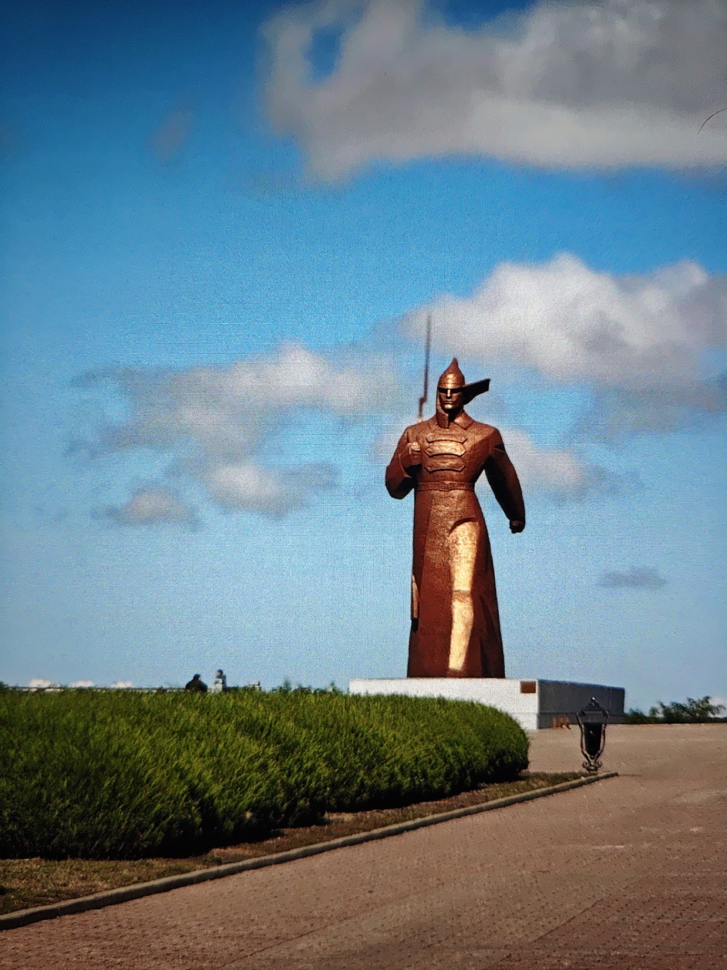 The image depicts a statue of a warrior, likely representing a historical figure, standing against a bright blue sky with scattered clouds. The statue is tall, made of a reddish-bronze material, and is depicted in armor, holding a sword aloft. It stands on a pedestal in an open area with a paved walkway in the foreground and a neatly trimmed green hedge to the left. In the background, there are a few indistinct figures and some modern buildings. This monument is the "Angel of Peace" located on the territory of the Stavropol Museum-Reserve "Russia – My History" in Stavropol, Russia.