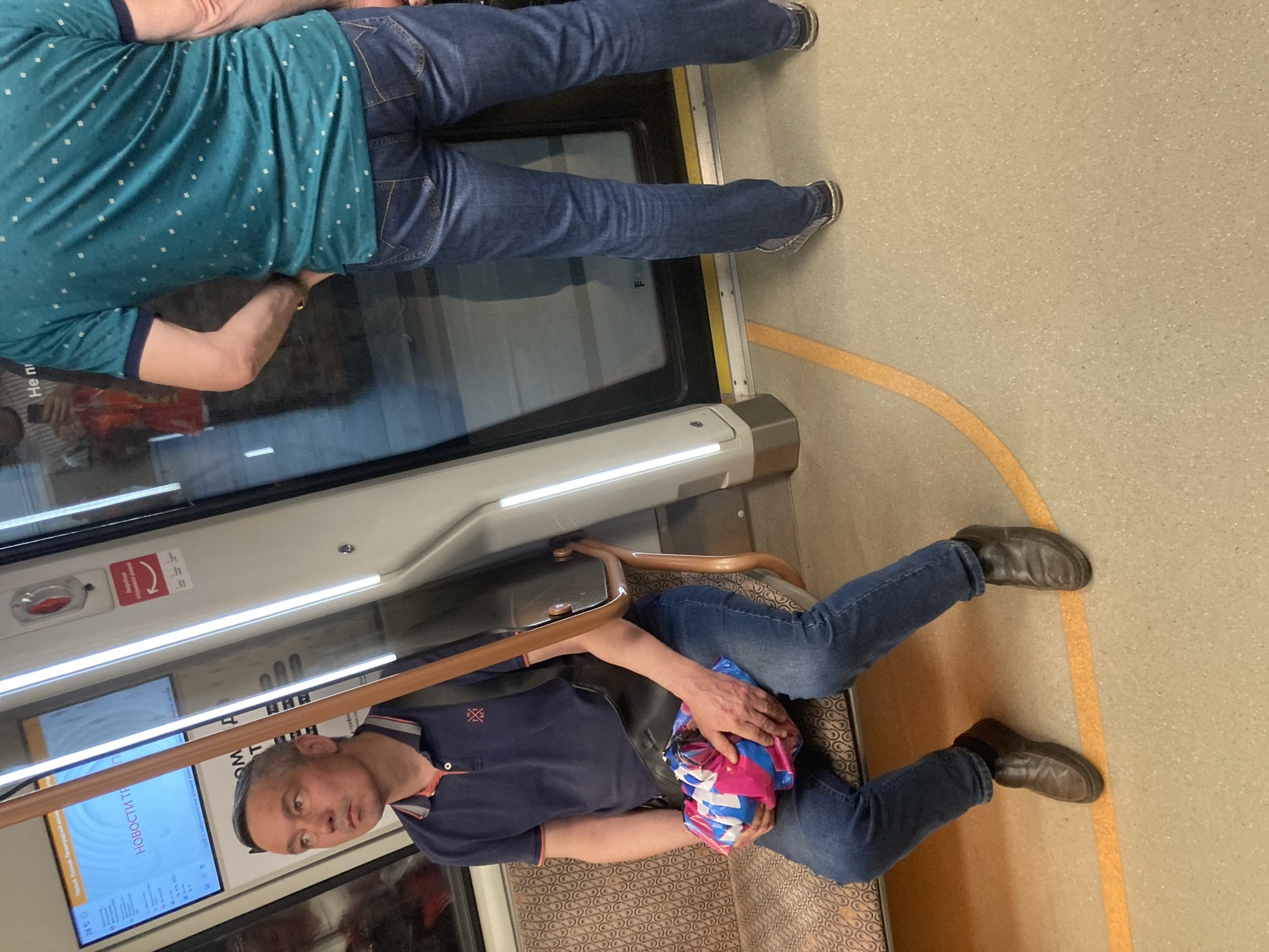 The image shows the interior of a train, likely in Moscow, Russia. A man wearing a blue polo shirt and jeans is seated, holding a colorful plastic bag in his lap. A second individual, seen from the waist up, is wearing a blue patterned shirt and jeans and is standing. The train car has seats with patterned upholstery, handrails, windows, and a digital screen displaying news or information. The floor is a light color with a curved yellow line. Text on the screen indicates that the location may be near the Leningradsky railway station.