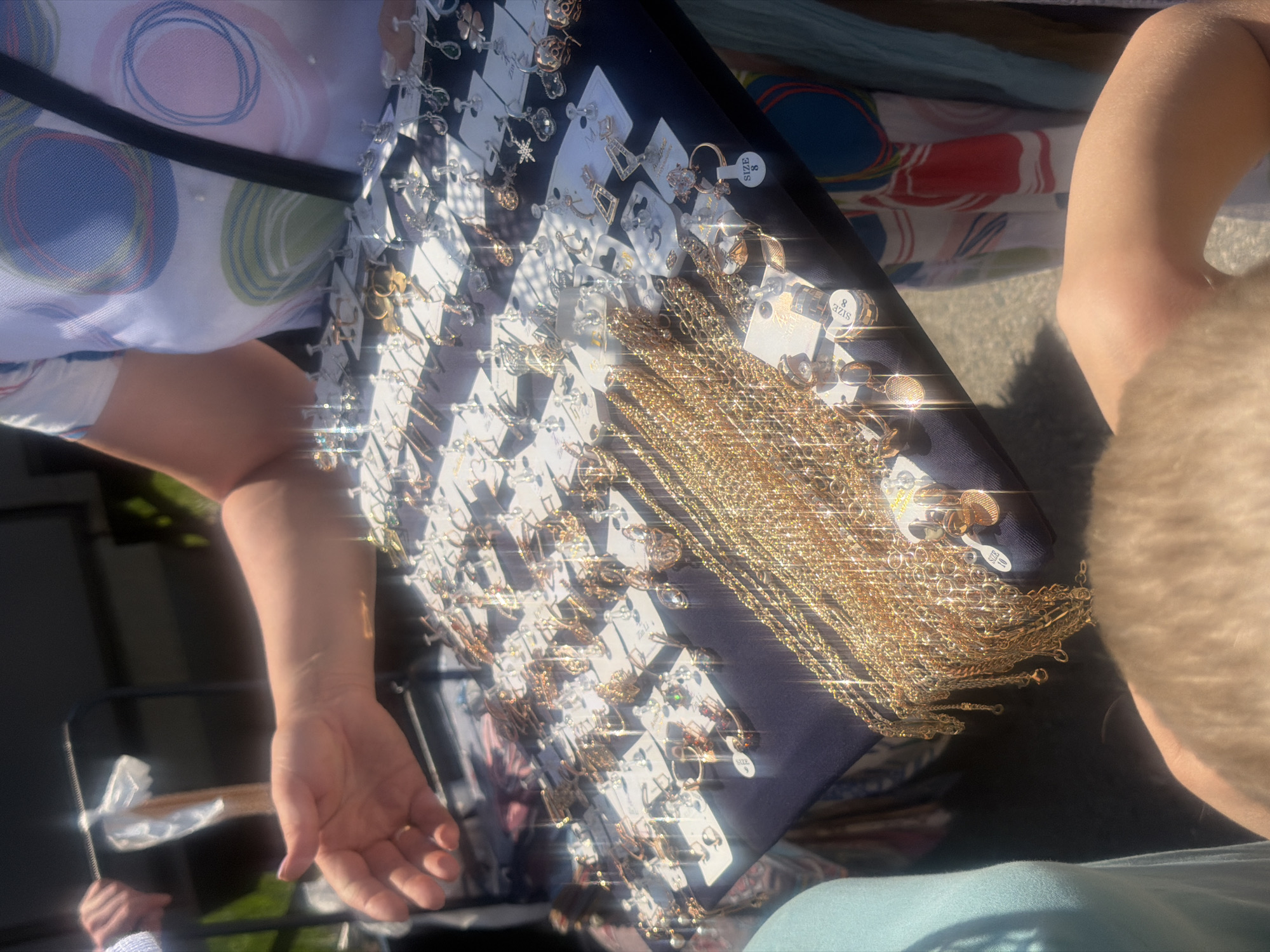 A person's arm extends into the frame from the left, fingers splayed, reaching towards a display of jewelry. The display is laid out on a dark fabric surface, covered in an array of necklaces and earrings. Many of the jewelry pieces are gold-colored and reflect the bright sunlight, creating a sparkling effect. Some earrings are attached to small white cards. In the background, a person wearing a white top with a colorful abstract pattern is partially visible, with a black strap across their chest. The scene appears to be outdoors, possibly at a market or street fair, with sunlight casting shadows and highlights. The location of Kanash, Russia, is not evident from the visual information in the image.