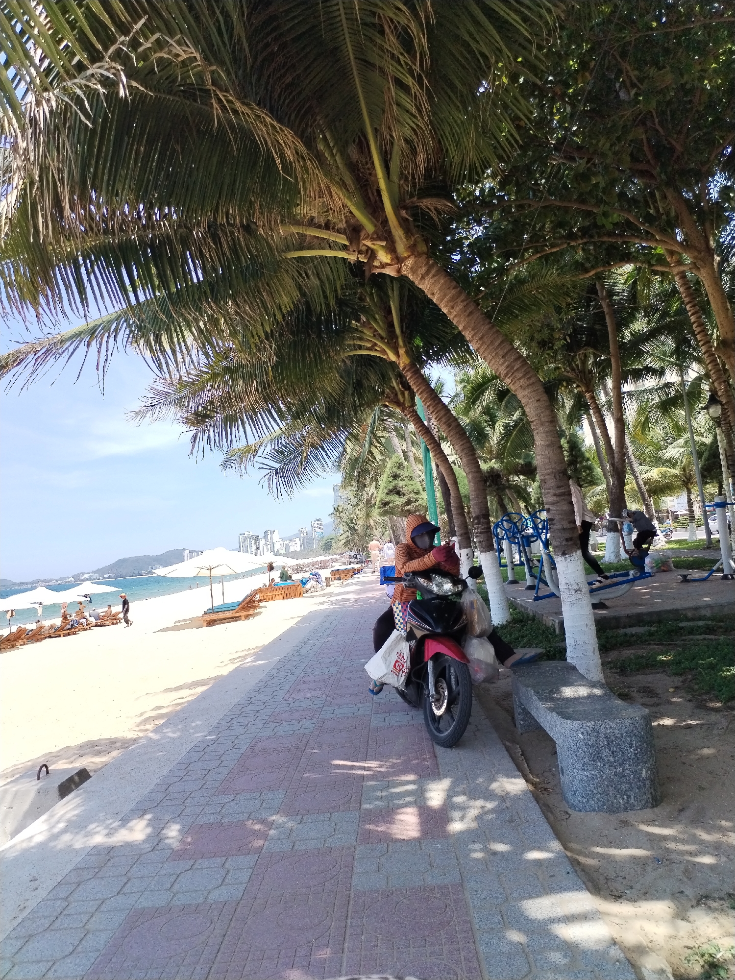 In Nha Trang Ward, Vietnam, a person is seated on a red and black motorcycle, wearing an orange hoodie and a mask. The motorcycle is parked on a tiled walkway adjacent to a sandy beach lined with lounge chairs and umbrellas. Behind the walkway, palm trees and other vegetation are visible, as well as outdoor fitness equipment and a stone bench. In the distance, the ocean stretches out towards a cityscape with tall buildings. The sky is clear and blue.