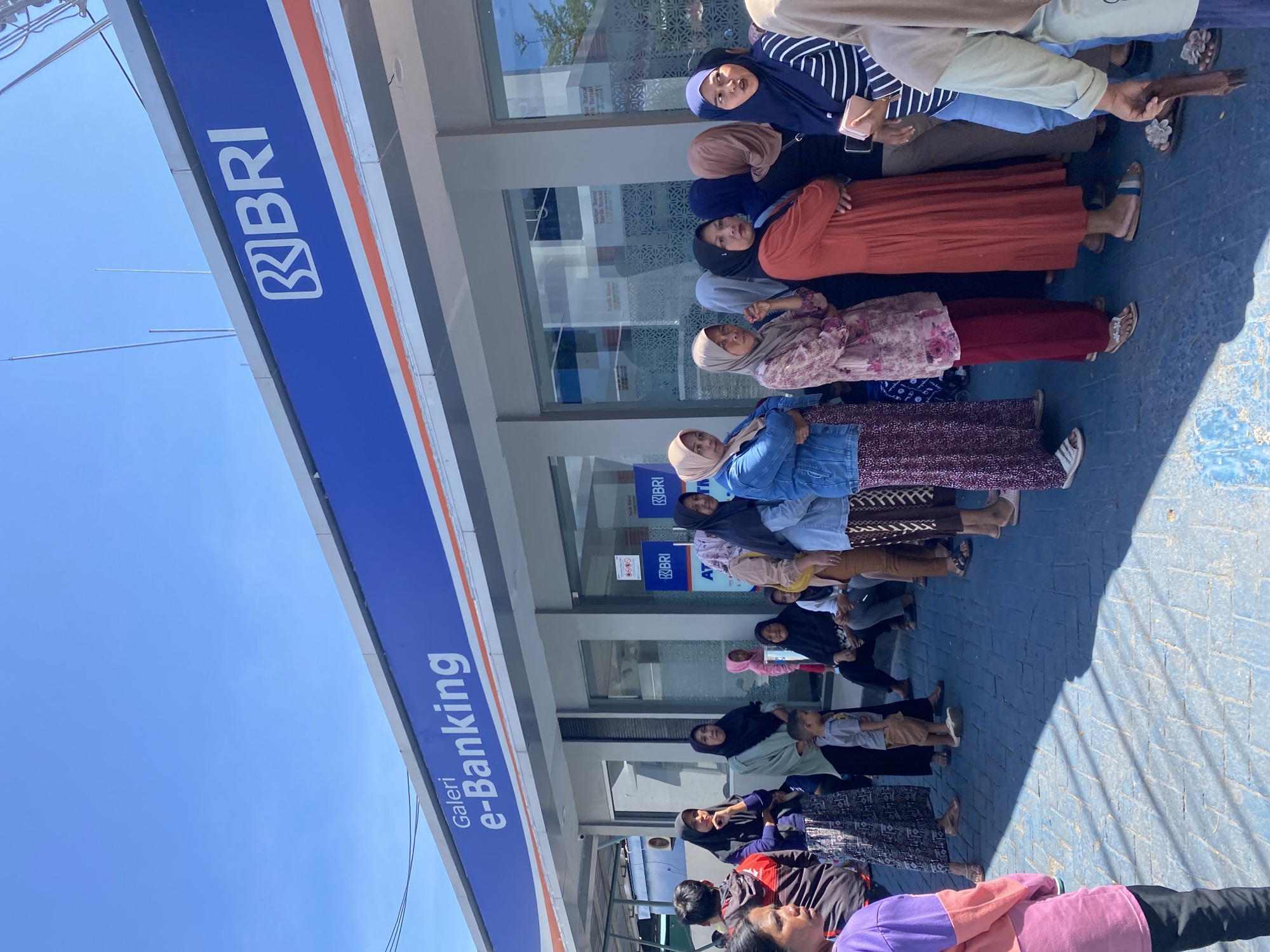 In Kelurahan Tappanjeng, Indonesia, under a clear blue sky, a line of individuals, predominantly women wearing headscarves, queues outside a BRI (Bank Rakyat Indonesia) e-banking gallery. The bank's facade features a prominent blue sign with white lettering that reads "BRI" and "Galeri e-Banking." The pavement in front of the building is a textured blue surface. The people in line are varied in age, with a young boy visible in the middle of the queue. Some individuals are looking forward, while others are engaged in conversation or looking down. The overall scene depicts a typical day at a banking institution in a localized Indonesian community.