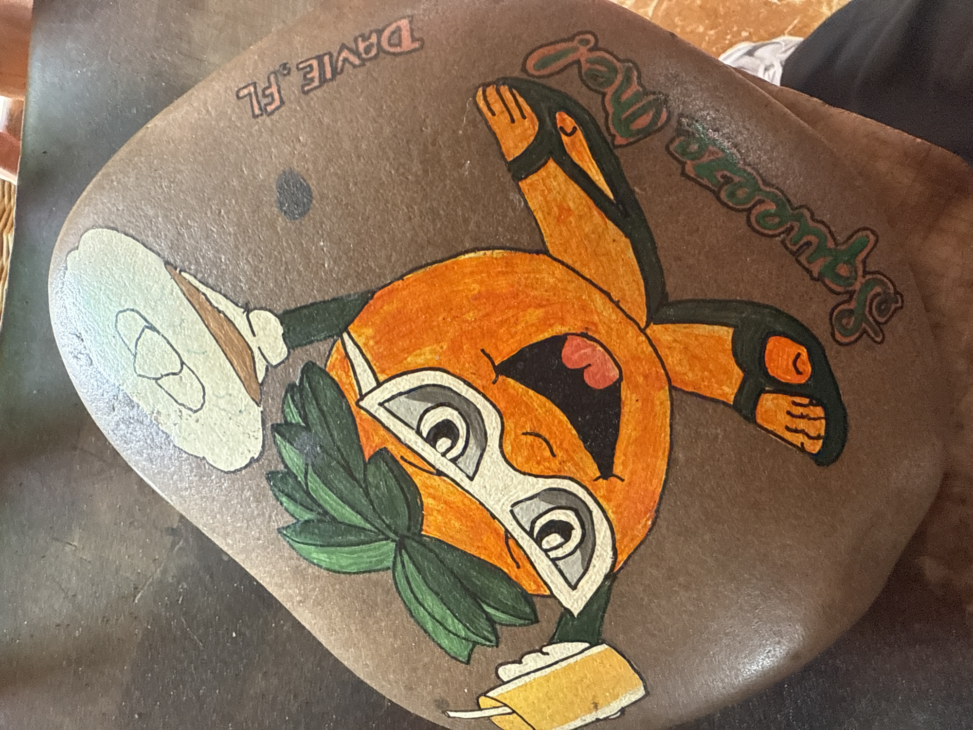 A rock painted with a cartoon orange character wearing sunglasses and holding a drink and a plate is depicted. The words "Squeeze Me!" and "Davie, FL" are also painted on the rock. The character appears to be associated with the city of Davie, Florida, United States of America.