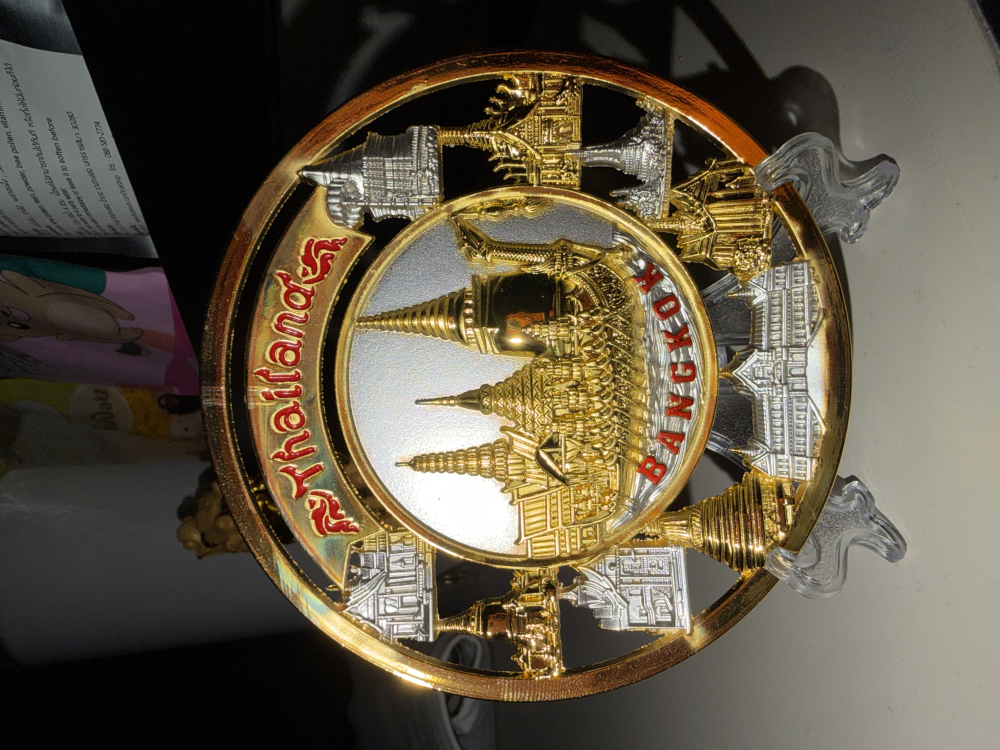 The image presents a circular, multi-layered metallic decorative plaque, likely a souvenir from Thailand, prominently featuring landmarks of Bangkok. The plaque measures approximately 20-25 cm in diameter and is supported upright by a clear, translucent plastic display stand.
The main subject is composed of gold and silver-toned metal with red enamel accents. The upper arc of the outer ring bears the word "Thailand" in stylized red script outlined in gold. The lower arc of the same ring displays "BANGKOK" in red block letters.
The central circular area of the plaque features a silver-toned background with raised golden architectural depictions. These include multiple tiered chedis (pagodas) and temple structures, characteristic of prominent Bangkok landmarks such as Wat Arun (Temple of Dawn) and components of the Grand Palace complex, including Wat Phra Kaeo (Temple of the Emerald Buddha). A golden royal barge is also depicted at the base of this central scene, representing an important cultural symbol associated with the Chao Phraya River in Bangkok.
The outer ring of the plaque, crafted from gold-toned metal, is designed with intricate, open-work silhouettes. These cut-out sections showcase additional Thai architectural elements, including both golden and silver-toned miniature stupas, pagodas, and traditional Thai buildings. The entire metallic surface is highly reflective, exhibiting prominent highlights from a direct light source.
The background behind the plaque is dark and out of focus. Faintly visible objects include a light-colored cylindrical container, a colorful, indistinct package, and a piece of paper with partially legible English and Thai text. The English text includes phrases like "protein, resin, powder, bee pollen, vitamin," and a web address "www.facebook.com/ukshop" along with a phone number "088-581-2774". No individuals are present in the scene.
