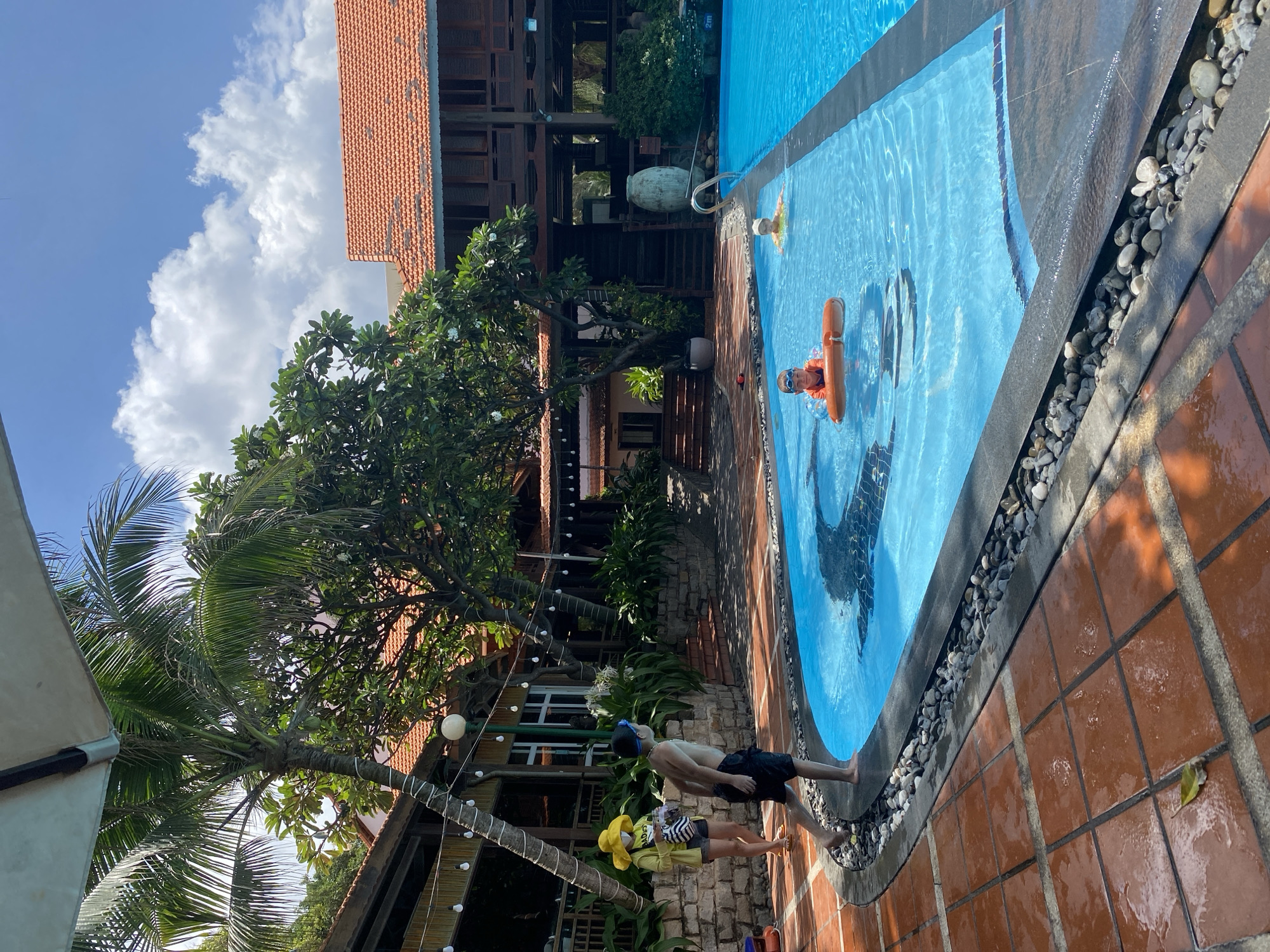 In a sun-drenched outdoor swimming pool area, a young boy, wearing swim trunks and goggles, is poised at the edge of the pool, ready to jump in. He is barefoot and appears to be in mid-motion, with one foot lifted. To his left, a young girl in a yellow hat, yellow cardigan, and shorts stands with a striped bag slung over her shoulder. She is also barefoot and looking towards the pool. In the background, a two-story building with a red tile roof and wooden balconies is visible. The pool itself is a bright blue, with a mosaic of a fish visible beneath the surface. Two other children are in the pool: one wearing a flotation device and goggles, and another further away, partially submerged. The area around the pool is paved with terracotta tiles, and a line of decorative pebbles borders the pool. Palm trees and lush greenery surround the scene, suggesting a tropical or subtropical climate. The sky is clear blue with fluffy white clouds. The location is identified as Phu Thuy Ward, Vietnam.