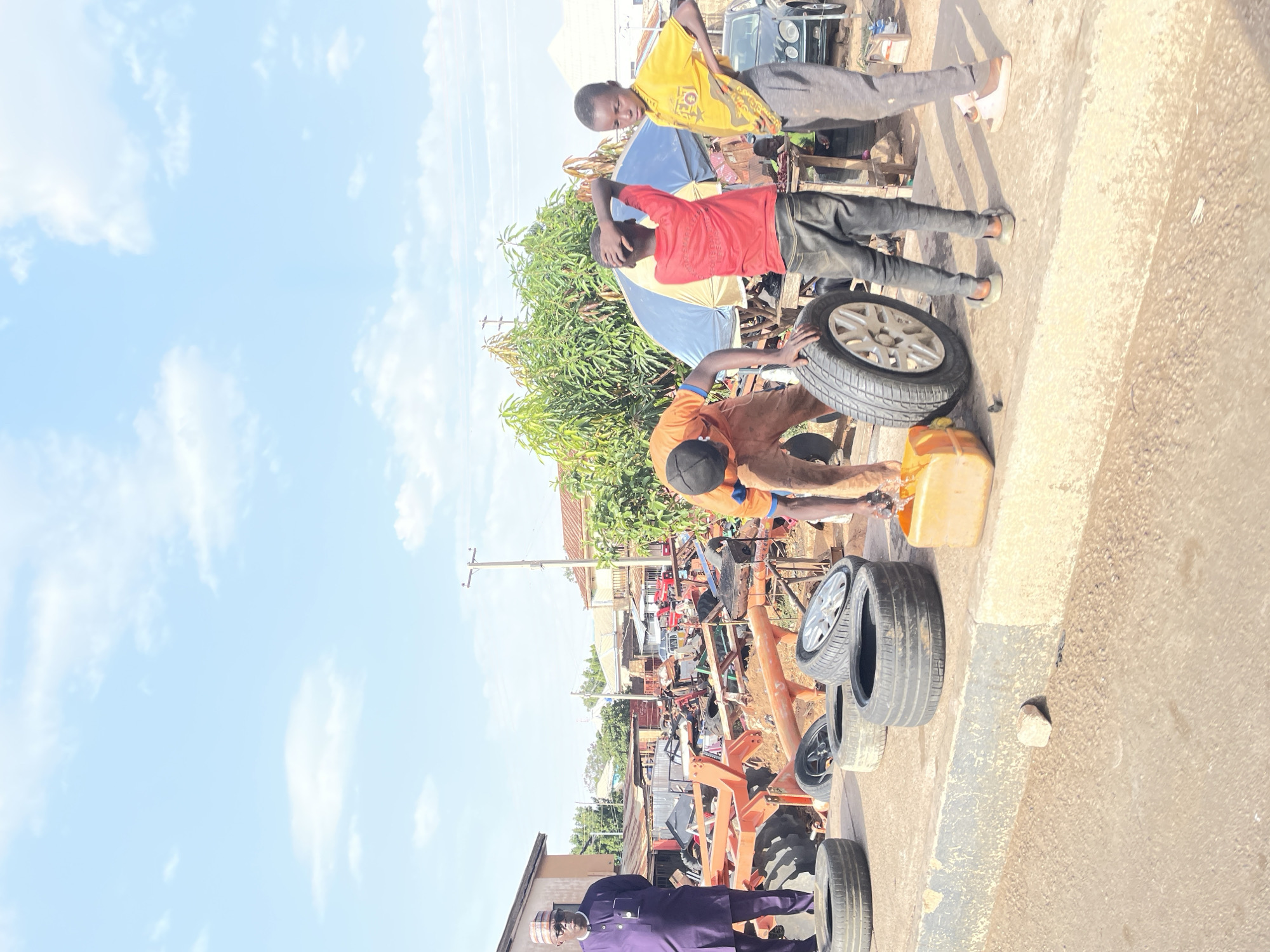 The image shows a roadside scene in Jos, Nigeria. Three men are visible at the forefront. One man is working on a tire with a yellow container beside him. Another man in a red shirt stands behind him, and a third man in a yellow shirt stands further back. Several tires are on the ground. In the background, a street scene is visible with a tractor, other vehicles, buildings, and vegetation. The sky is blue with scattered clouds.