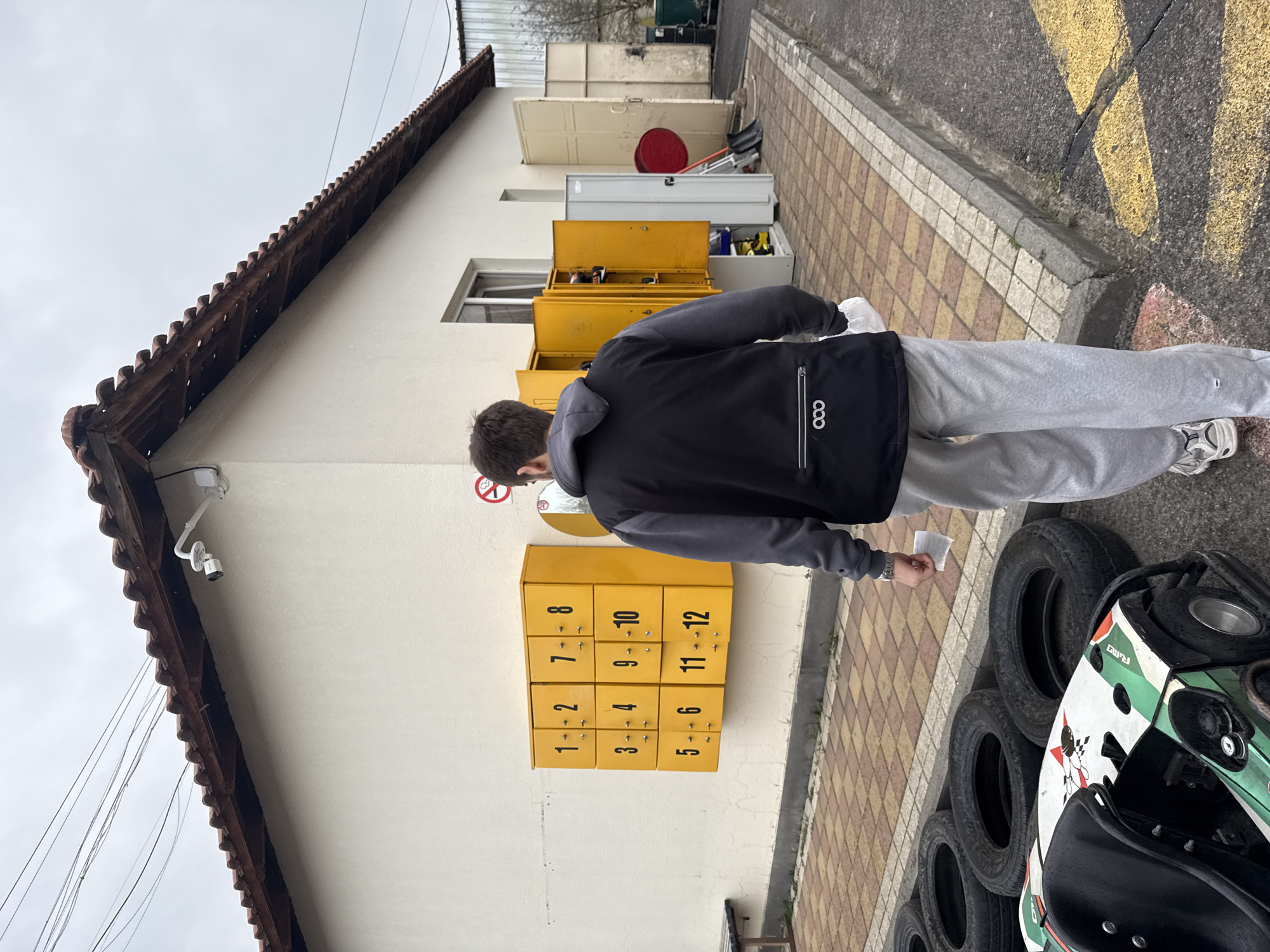 A medium shot depicts a young man, seen from behind, standing beside a building and a go-kart. He wears a black and gray jacket and gray sweatpants and holds a small piece of paper in his hand. He is standing next to a row of yellow numbered storage boxes attached to the building's wall. The boxes are labeled with numbers ranging from 1 to 12. The building is cream-colored with a brown tile roof and is equipped with a security camera. Several tires are stacked next to the go-kart. The location's exact city or country is not discernable from the image provided, as the image contains no clues to confirm the location as Yerevan, Armenia. The scene is outdoors on a paved surface.