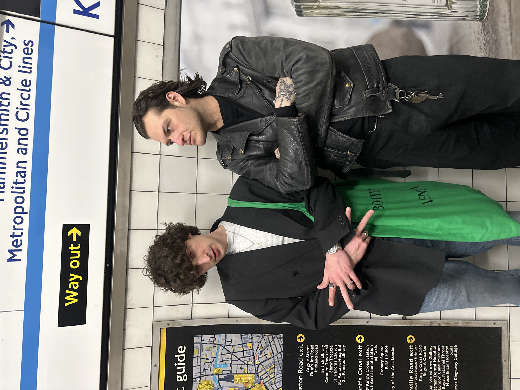 Two males stand against a white tiled wall in the King's Cross St. Pancras Underground station, London, United Kingdom. Above them, a white sign indicates "Hammersmith & City, Metropolitan and Circle lines" and a "Way out" sign with a right-pointing arrow. To the left, a black information board with a yellow border displays a local map and lists local exits such as "Euston Road exit" and "Regent's Canal exit," confirming the station as King's Cross.
The male on the left, with dark curly hair, wears a grey crew-neck t-shirt, an oversized black blazer, and light blue denim jeans. He has a silver-colored rectangular wristwatch on his left wrist and multiple silver-colored rings on both hands. He carries a bright green tote bag, partially displaying "BOTTEGA VENETA" text, over his right shoulder. He faces the camera, exhibiting a slight smirk and making a two-handed gesture with splayed fingers.
The male on the right, with dark wavy hair styled in a mullet, wears a black t-shirt, a distressed black leather motorcycle jacket with brass-colored studs and zippers, and black trousers. Keys are attached to a black leather strap hanging from his right belt loop. A tattoo is visible on the back of his left hand. He stands with his arms crossed, looking to his left, away from the camera. A partial view of a ribbed metallic cylindrical object is visible on the far right of the frame.