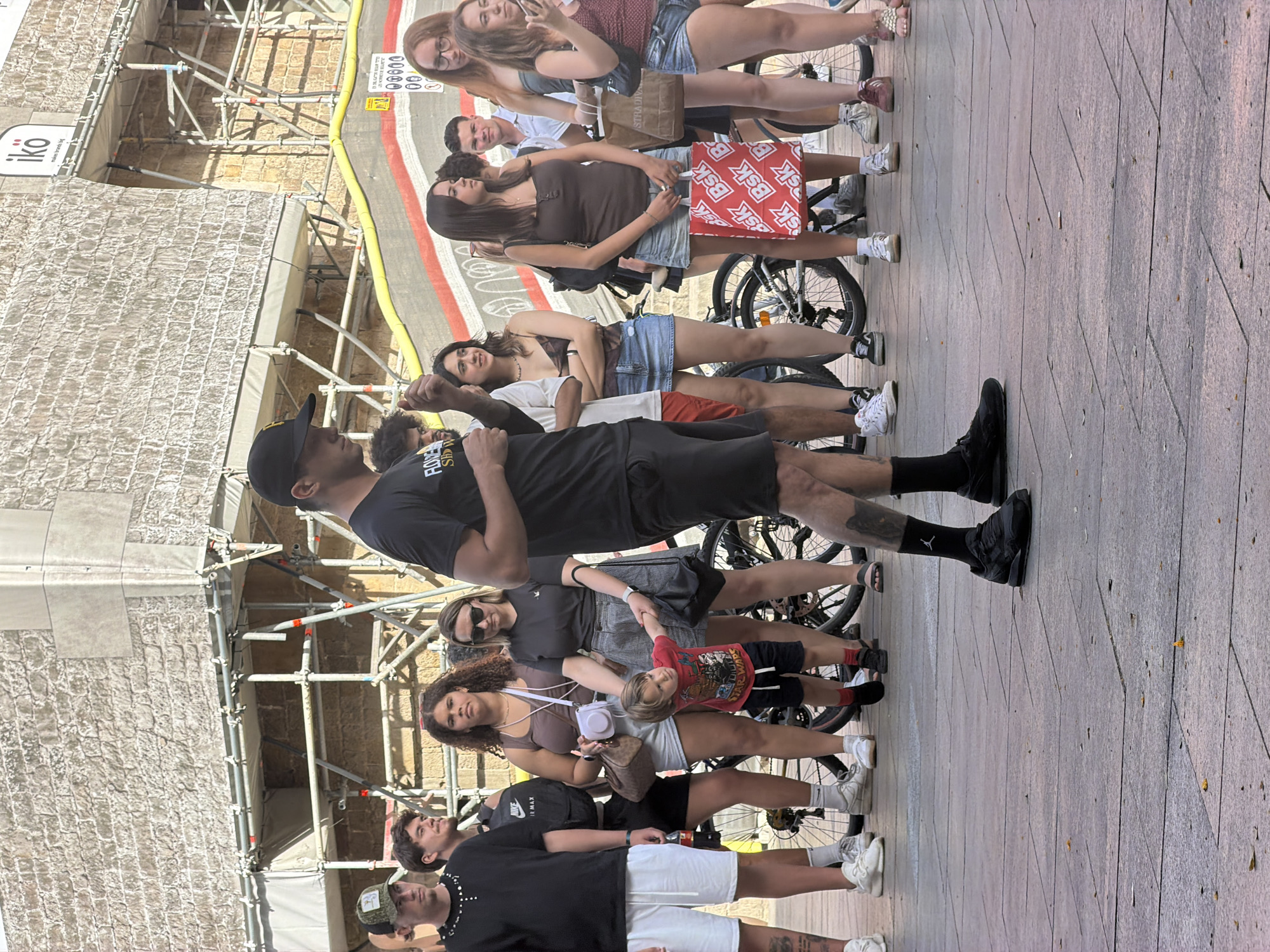 A group of people are gathered outdoors in what appears to be a public square. The architecture in the background, with its stone facade and scaffolding, suggests a historical or undergoing renovation setting. In the foreground, several individuals are standing, including a man in a black t-shirt and black baseball cap who is facing away from the camera, and a young child in a red t-shirt is holding his hand. Other people in the group are dressed in casual summer attire. Bicycles are visible in the background, indicating a mode of transport for some. The ground is paved with large stone tiles. There are commercial elements visible, such as a sign with the word "iko". The overall impression is a casual gathering in an urban environment. Based on the scaffolding and the style of architecture, the scene could plausibly be in Barcelona, Spain, possibly near a construction site or a landmark undergoing preservation.