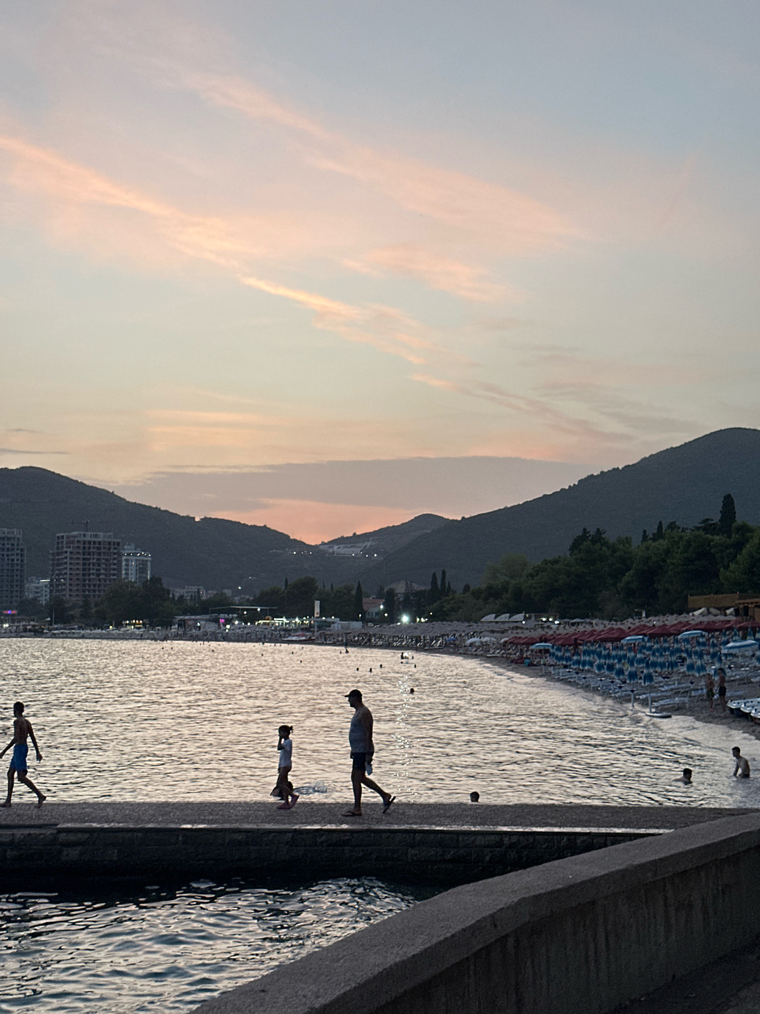 This is a horizontal, eye-level, medium shot of a beach scene at dusk in Budva, Montenegro, likely near Slovenska Plaza given the architectural style of the buildings and the mountainous backdrop. The sky is a soft blend of pale blue and pink from the setting sun, with wispy clouds. In the foreground, a dark stone pier extends into the water, where three figures are walking: a young boy in blue shorts on the left, a young girl in a white top and pink shorts in the middle, and a man in a gray t-shirt and dark shorts on the right. Their silhouettes are cast against the shimmering water.
The water is calm with gentle ripples reflecting the sky's colors. To the right of the pier, a crowded beach is visible, lined with numerous blue and white striped lounge chairs and red umbrellas. People are scattered on the beach and in the shallow water, some swimming. In the middle ground, a line of trees separates the beach from a town with several modern, high-rise buildings, some appearing to be under construction. Beyond the town, dark, green, tree-covered mountains rise, forming a dramatic silhouette against the dusky sky. The overall mood is serene and tranquil, capturing the end of a summer day.