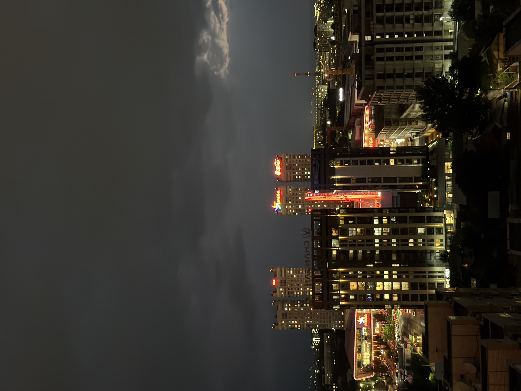 The image shows a night view of Almaty, Kazakhstan. Tall buildings are visible, illuminated by numerous windows. The sky is dark with some cloud cover. Several buildings display bright, colorful signage, including recognizable logos. The city skyline stretches into the distance, marked by various lights and structures. The perspective suggests the photo was taken from a high vantage point.