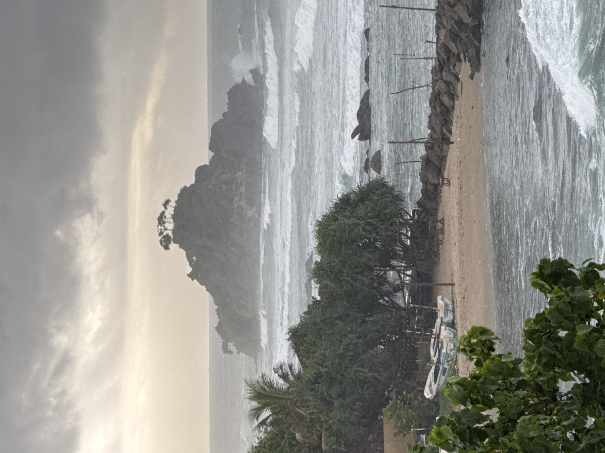 An eye-level, medium shot captures a coastal scene in Ahangama, Sri Lanka, under a hazy, overcast sky. The ocean is active with moderately sized waves crashing against a rocky shoreline. In the middle ground, a small, tree-covered island rises from the sea. Foreground elements include lush green foliage on the left, a sandy beach with two weathered fishing boats, and a breakwater constructed of rocks. The overall atmosphere is moody and somewhat dramatic due to the weather.