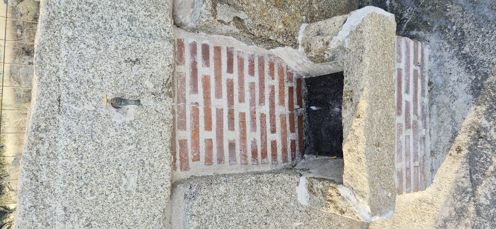 This is a close-up, vertical, outdoor, eye-level shot of a water fountain. The fountain is constructed from large, rough-hewn granite blocks and red bricks. A brass faucet protrudes from the granite at the top center of the frame, and a stream of clear water is flowing from it. The water falls into a basin lined with a black grate. The background consists of dry vegetation and a metal fence, suggesting a rural or arid environment. The lighting indicates it is daytime, likely late afternoon due to the shadows.