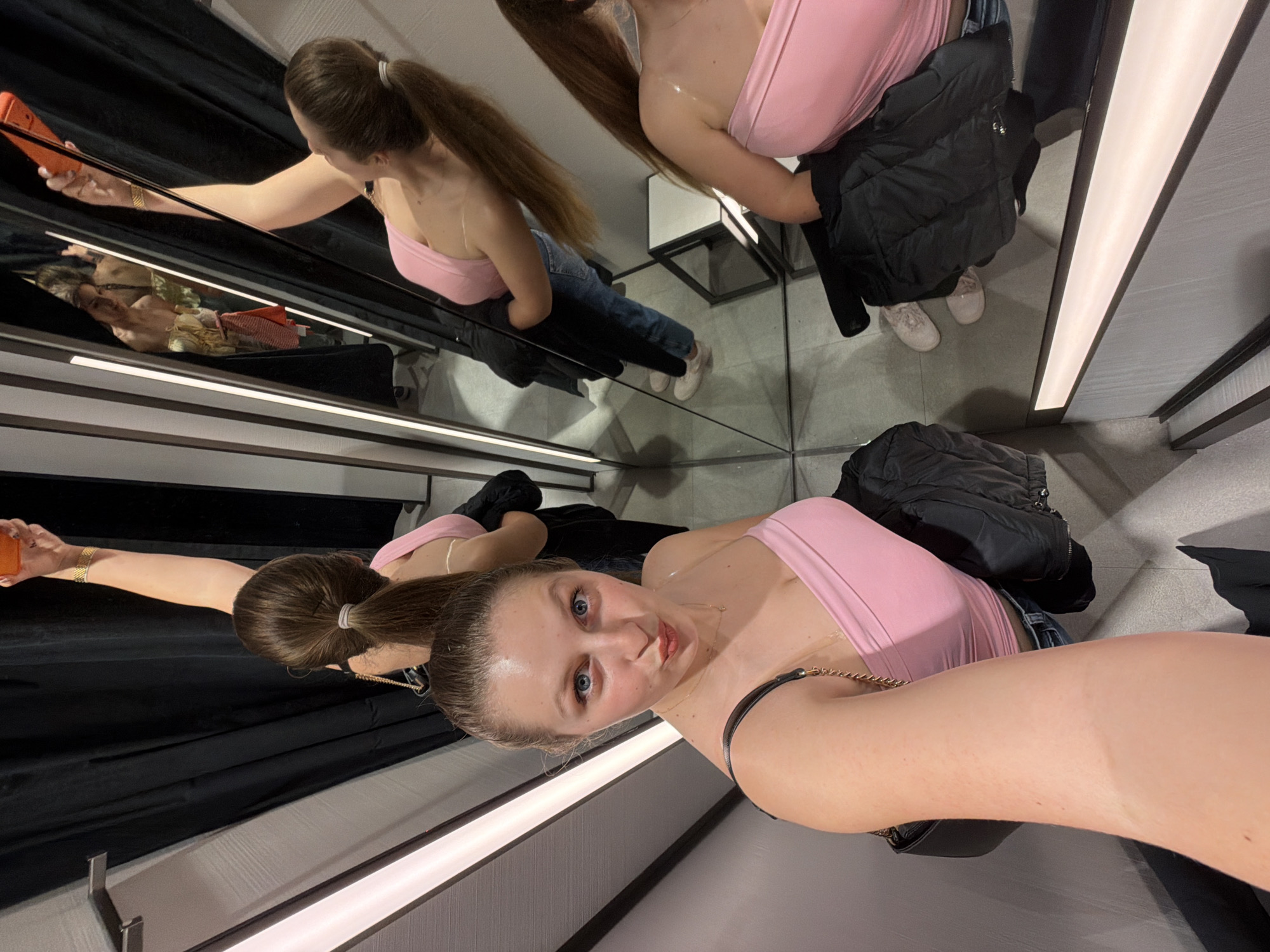 The image depicts a young woman taking a mirror selfie inside a dressing room, likely in a retail store in Burgas, Bulgaria. The setting is characterized by multiple mirrors that create numerous reflections of the subject and the interior.
The main subject is a woman with long brown hair, tied back in a ponytail. She is wearing a pink strapless or bandeau-style top, blue jeans (visible in reflections), and light-colored sneakers. A black puffer jacket or vest is draped over her arm, and she carries a black handbag with a gold chain strap. In one of the reflections, she is seen holding a smartphone with an orange case in her outstretched hand, indicating she is actively taking the selfie.
Her face is visible in the foreground, showing her with blue eyes looking directly at the camera, a slight pout on her lips, suggesting a confident and playful expression. The dressing room features black curtains, which provide privacy, and integrated strip lighting runs along the base of the mirrored walls, illuminating the space. The fragmented reflections create a dynamic and multi-angled view of the woman and her attire. No visible text appears in the image.
