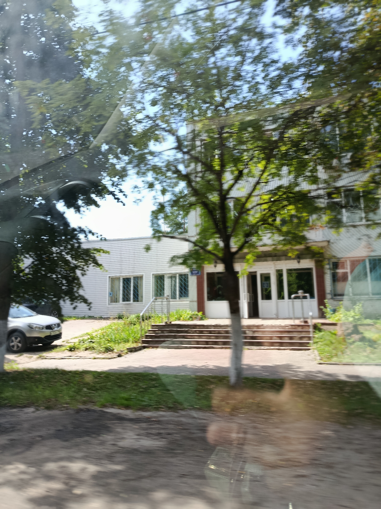 The image shows a building with a light-colored facade and several windows. In front of the building, there is a set of stairs leading up to a double door entrance. To the left of the entrance, a silver car is parked. The area is surrounded by trees and grass, suggesting a daytime setting. The image is slightly blurred, indicating movement.