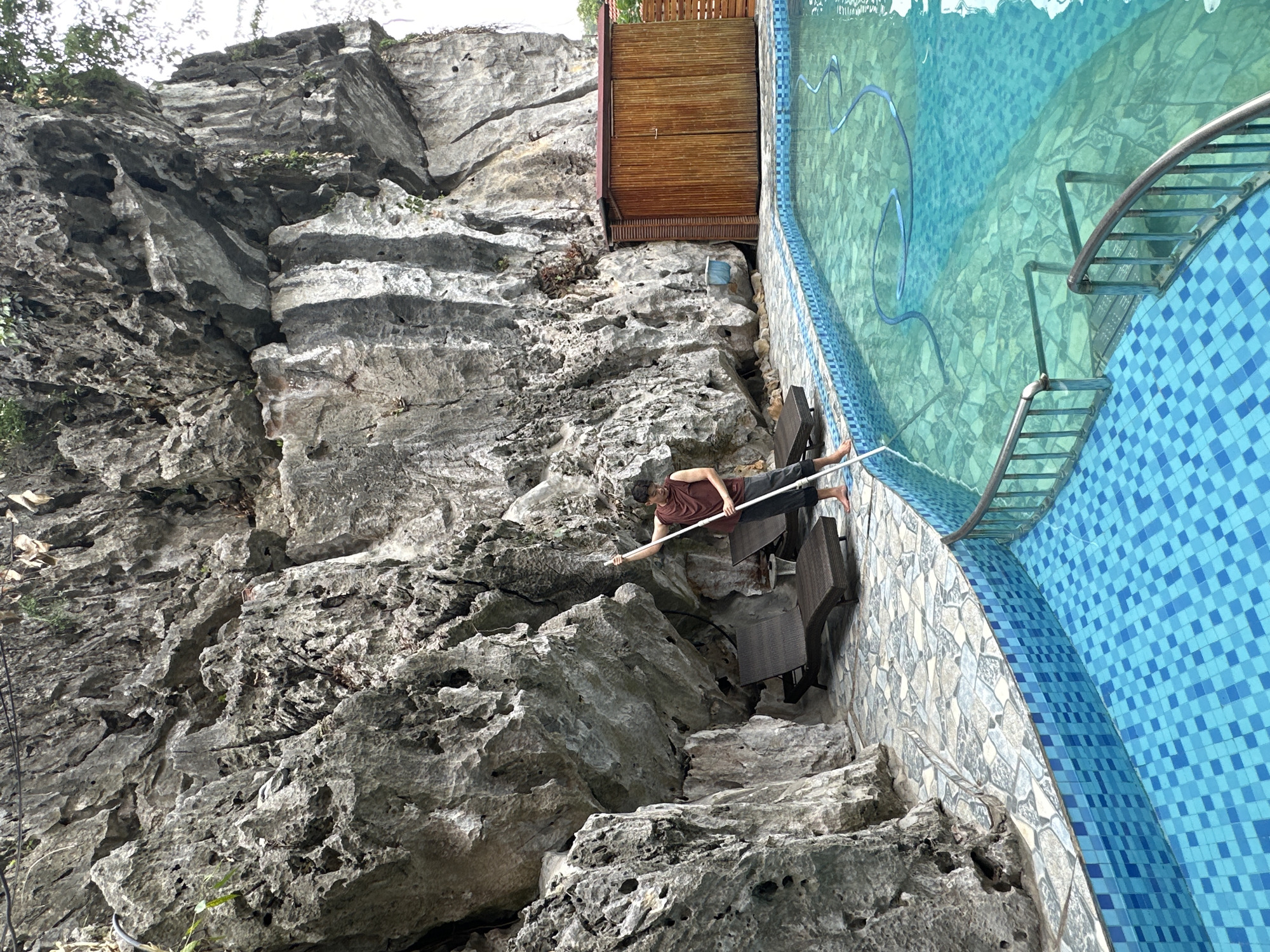 The image depicts an outdoor swimming pool adjacent to a large, rocky cliff face. A young man, wearing a maroon tank top and dark pants, stands on the tiled pool deck and uses a long-handled pool cleaning tool to skim the surface of the clear turquoise water. Two lounge chairs are visible on the stone-tiled area between the pool and the cliff. To the right of the cliff, a small wooden shed with a reddish-brown roof is situated. The overall scene suggests a private residence or resort in a natural, mountainous environment, possibly within Nam Hoa Lư Ward, Vietnam, given the geological formations common to the region.