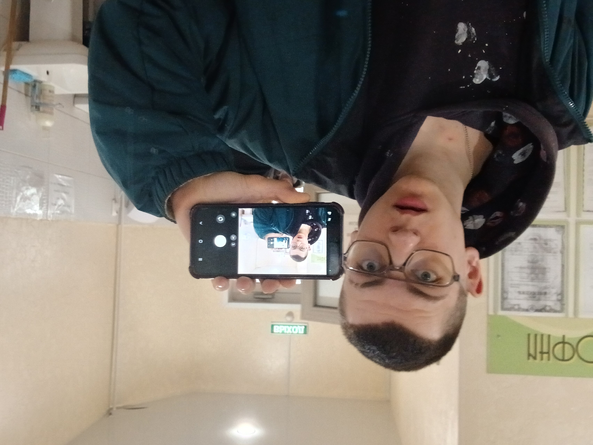 A person with short dark hair and glasses is holding a smartphone to take a picture of himself. The smartphone screen shows a mirrored image of the same person. He is wearing a dark jacket and a dark hooded sweatshirt with paint splatter. The background appears to be an interior hallway of a building. A green sign with Cyrillic letters reading "ВЫХОД" (Exit) is visible above a doorway. Other signs and posters are on the wall, also with Cyrillic writing. The location is suggested as Gory, Russia based on the language used on the signs.