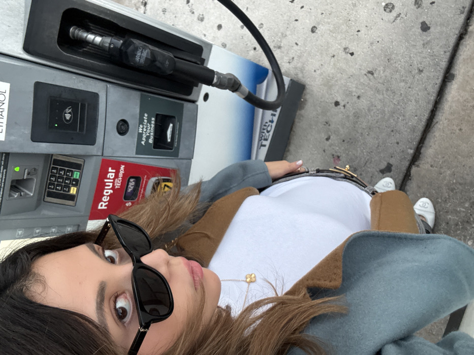 A woman takes a selfie at a gas station. She wears sunglasses, a white top, and a coat with brown and light blue accents. Her necklace is visible. The gas pump is to her left, with a nozzle inserted and displaying a "Regular" price of $4.269 with "TECHRON". Text on the pump reads "We Appreciate Your Business". She also wears white shoes.