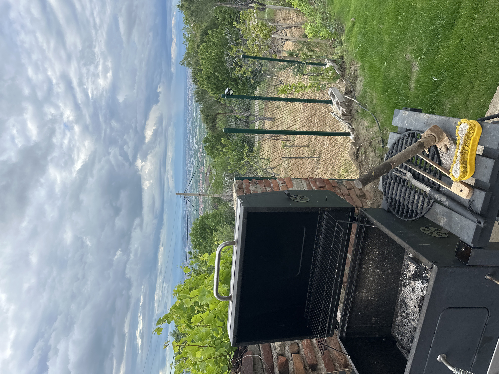 The image captures a scene in Brestnik, Bulgaria, featuring a weathered black charcoal grill positioned on a brick base. The grill's lid is open, revealing remnants of ash and charcoal inside. A metal grill and various grilling utensils, including a wooden-handled scraper with a yellow brush, rest atop the grill. The environment is rural, with green foliage and a vineyard visible. A green metal fence separates the foreground from a sloping landscape. In the distance, a cityscape stretches towards a body of water under a sky filled with dramatic, cloud-laden formations.