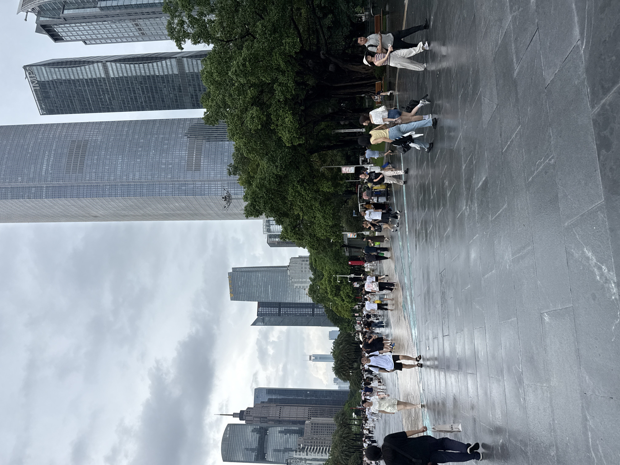 The image depicts a bustling pedestrian plaza in the Tianhe District of Guangzhou, China, characterized by a cloudy sky and a wet, stone-tiled ground, suggesting recent rain. A diverse crowd of people, dressed in casual summer attire, walk across the plaza in various directions. In the background, towering modern skyscrapers with glass facades dominate the skyline, indicative of a major urban commercial and business hub. Lush green trees are interspersed between the buildings, adding a touch of nature to the concrete jungle. The composition captures a moment of everyday urban life, with individuals and small groups engaged in walking, shopping, and socializing. The wet ground reflects the overcast sky and the surrounding architecture.