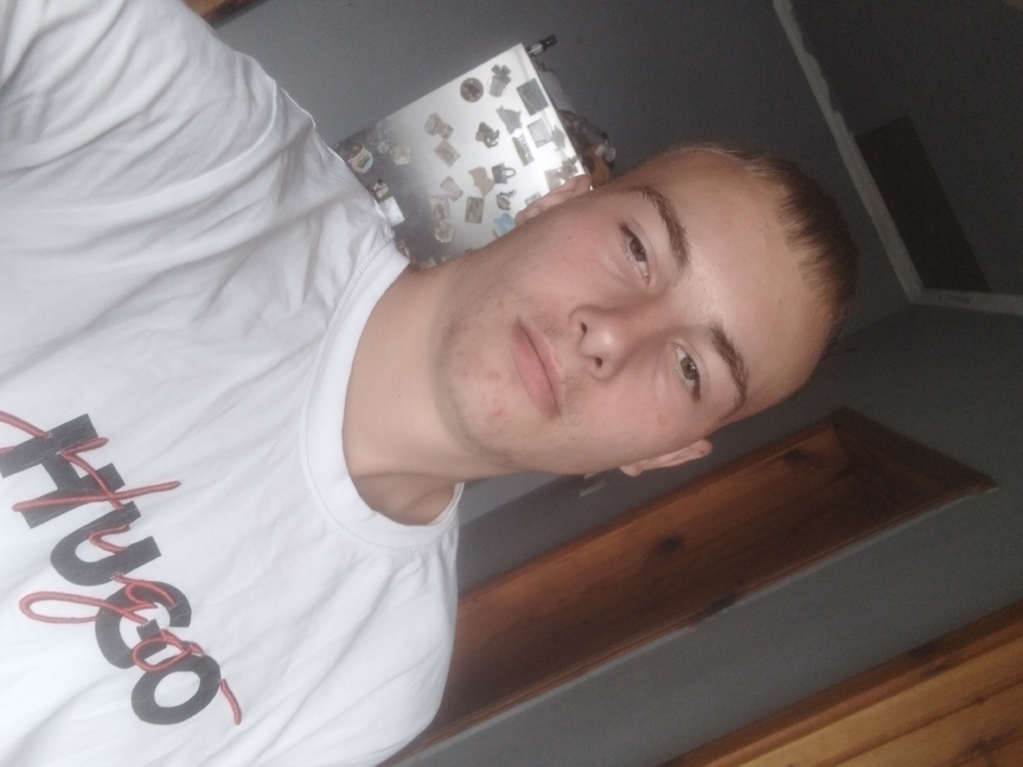 The image shows a selfie of a young man in his late teens or early twenties, wearing a white t-shirt with "HUGO" printed on it. He has short, light brown hair and light brown eyes. The background appears to be an indoor setting, possibly a room in Plastina, Bulgaria, with a grey wall and a wooden door or frame. A refrigerator with several magnets is visible on the left side of the frame. The lighting is somewhat dim, casting shadows and creating a slightly grainy effect.