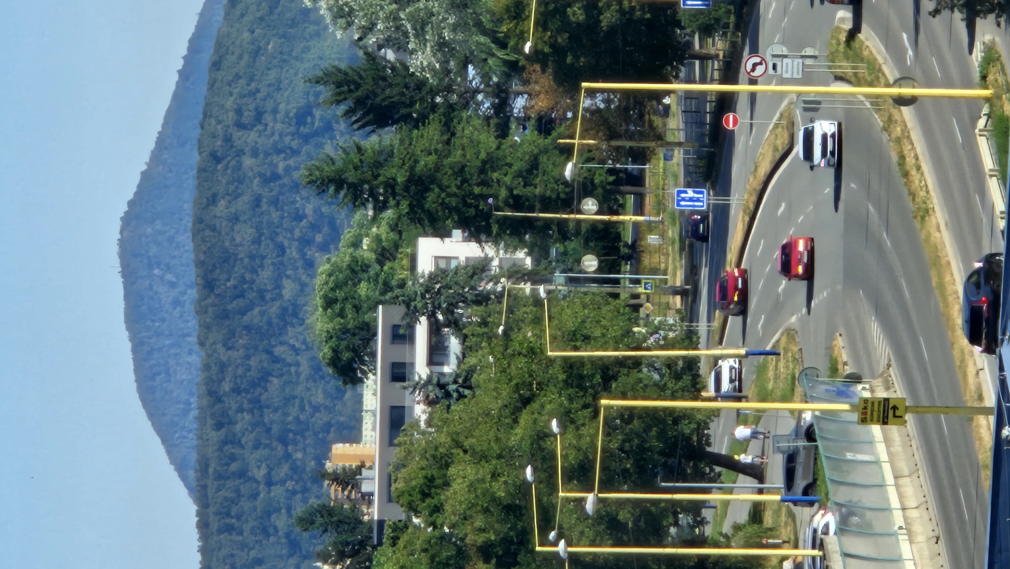 This is an aerial photograph of a city street in Prešov, Slovakia, on a clear, sunny day. The foreground shows a multi-lane road with several cars driving. A dark SUV is parked in the lower center, and a red sedan and two white cars are visible on the road. Streetlights with yellow poles line the road. In the middle ground, lush green trees and a modern white building are visible. A small group of people are walking on the sidewalk. The background features a densely forested mountain range and a clear blue sky.